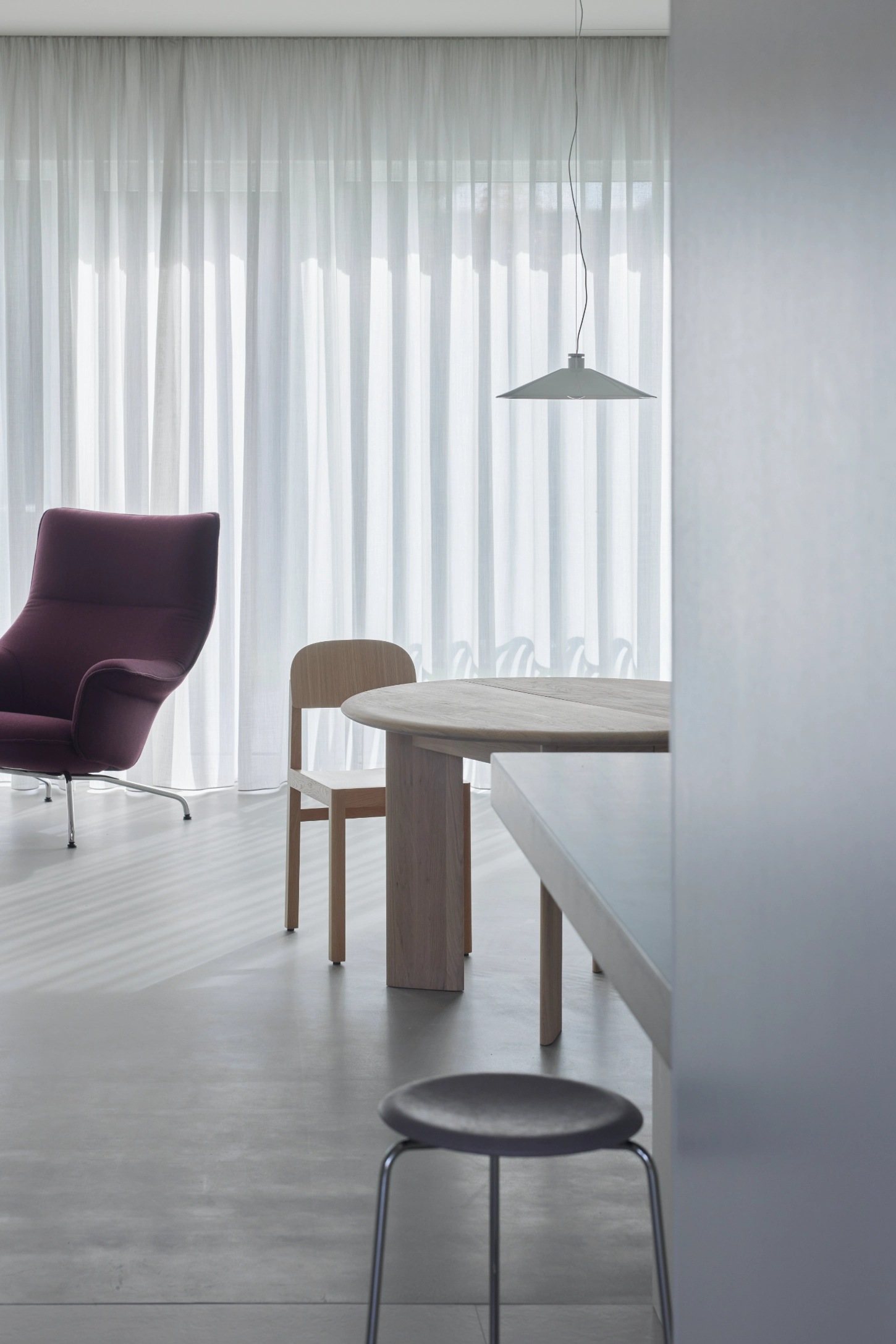 VVV by i.B Archstudio — A minimalist interior scene featuring a light timber circular dining table with matching chair, positioned beneath a suspended graphite-grey pendant lamp.