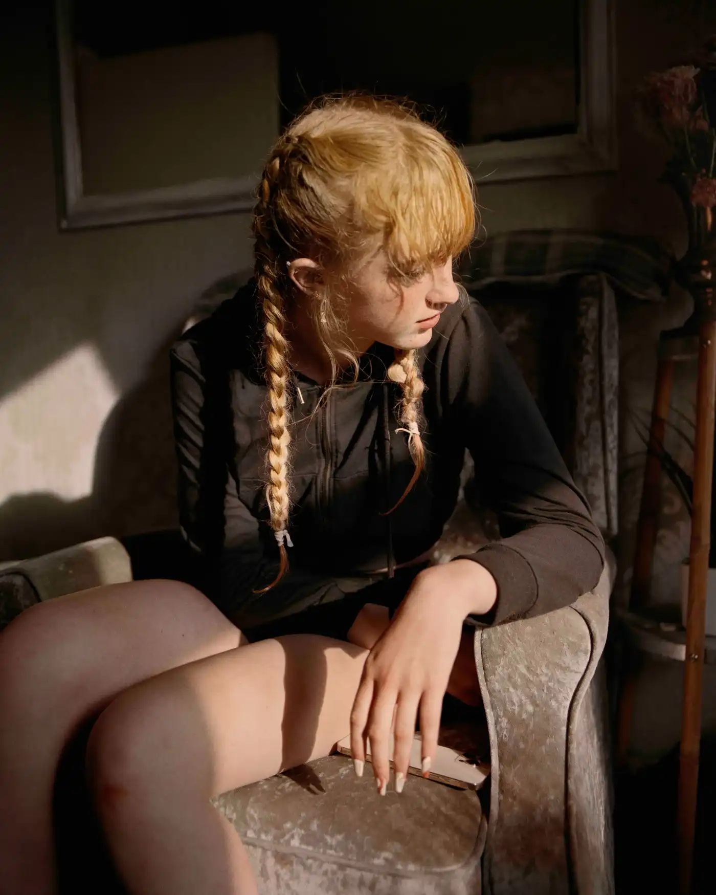 Land Loss by Max Miechowski — A figure with blonde hair in two braids sits in profile on an upholstered chair, wearing a black long-sleeved top.