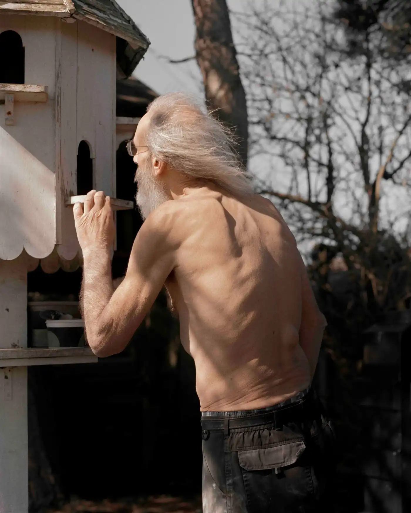 Land Loss by Max Miechowski — A shirtless man with long grey hair and beard examines a wooden birdhouse structure mounted on a white-rendered wall.