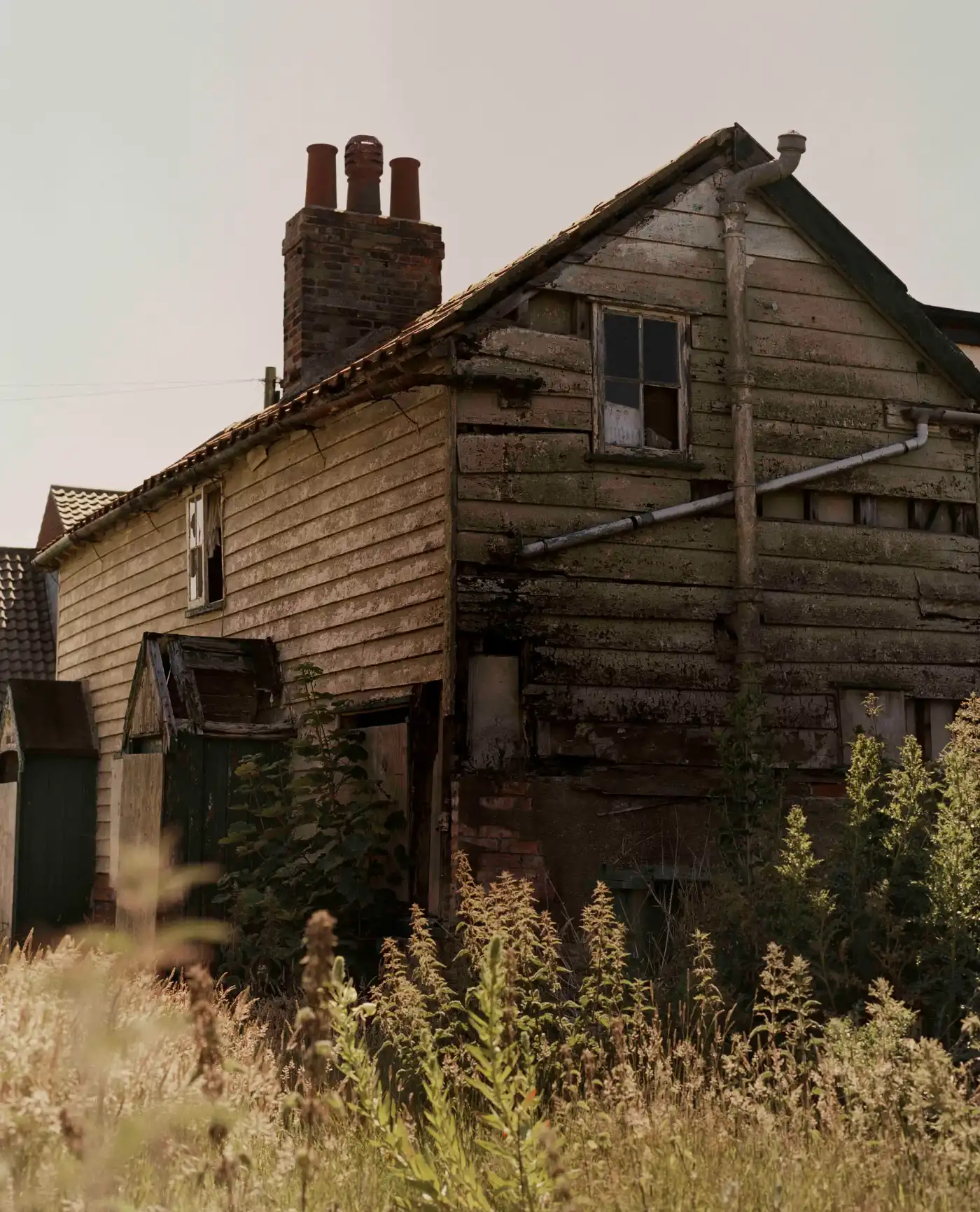 Land Loss by Max Miechowski — A timber-clad residential structure sits precariously at the cliff edge, its wooden siding weathered to a silvered grey-brown patina.