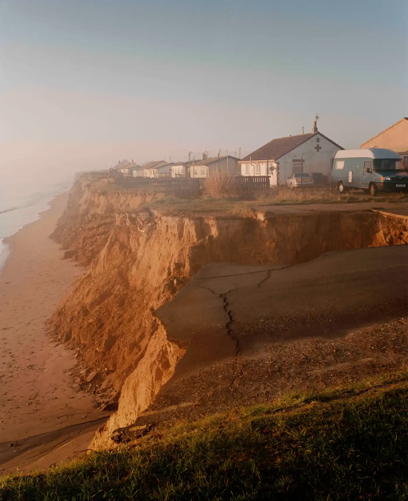 Land Loss by Max Miechowski — A row of residential structures perches precariously at the edge of a eroding sandstone cliff, their rear facades aligned with the cliff edge and minimal...