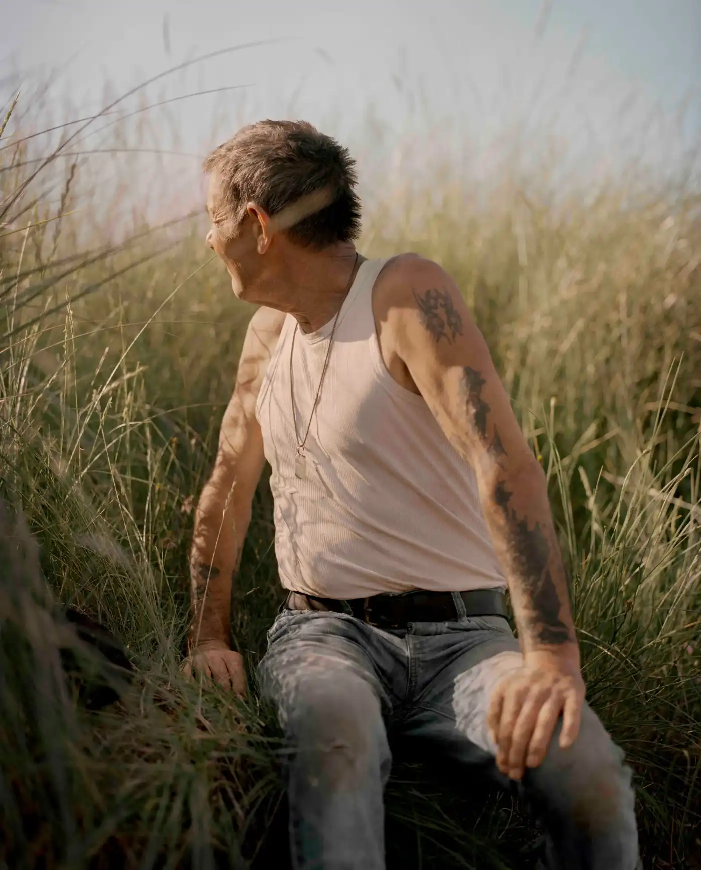 Land Loss by Max Miechowski — A man in a white sleeveless tank top and grey jeans sits crouched among tall, dry grasses in what appears to be coastal marshland.