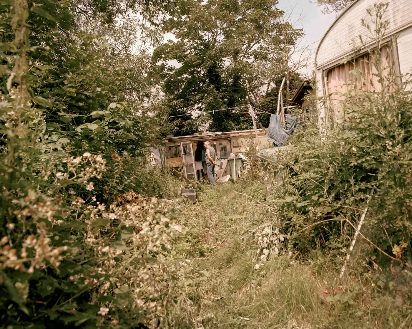 Land Loss by Max Miechowski — A modest single-story structure sits precariously at the cliff edge, its cream-rendered walls and pitched roof visible through encroaching wild vegetation.