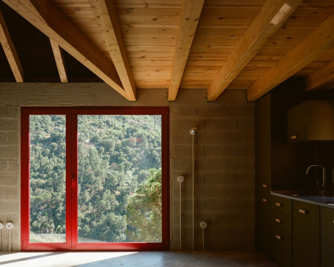 House in Ancede by atelier local — A bathroom threshold framed by a bold crimson-red metal door and window assembly that opens onto dense forest vegetation.