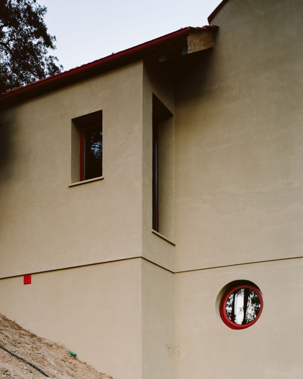 House in Ancede by atelier local — The corner elevation reveals the project's material and compositional strategy: cream-rendered masonry walls meet a burgundy metal roof trim, establishing a...
