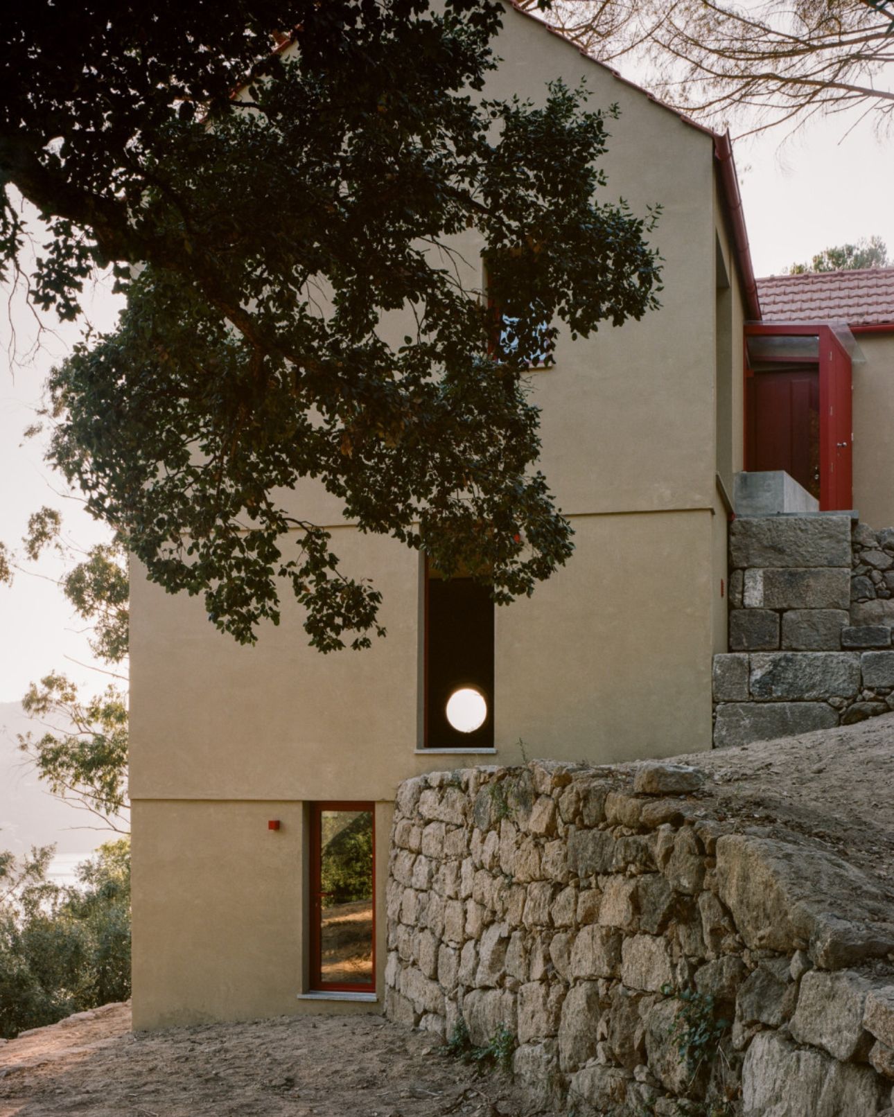 House in Ancede by atelier local — A rear elevation view of the concrete-rendered addition reveals its stacked volumetric composition against the existing stone masonry wall.