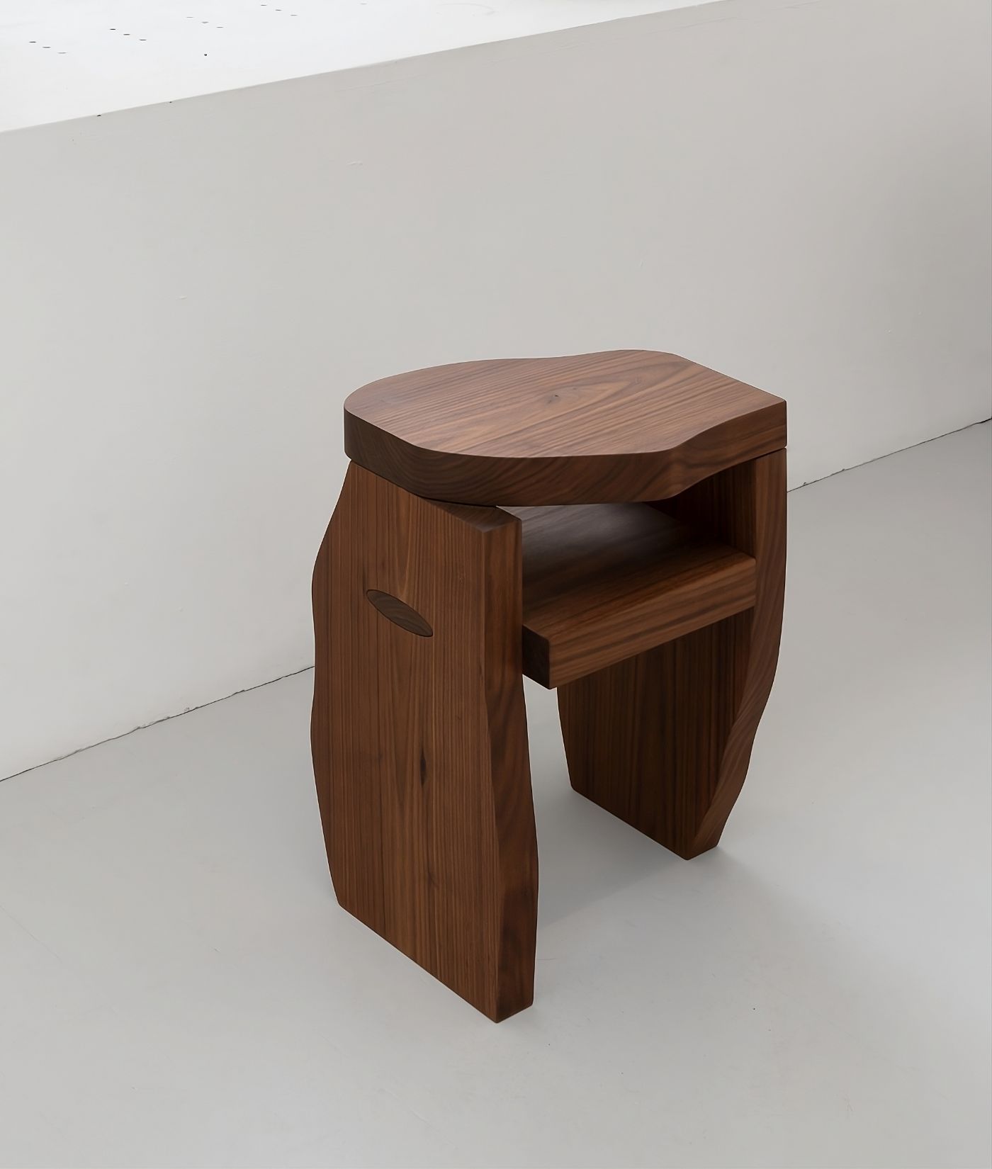 Selected Works by Bahraini-Danish — A walnut side table with sculptural proportions features an undulating tabletop with softly rounded edges that overhang two curved leg supports.