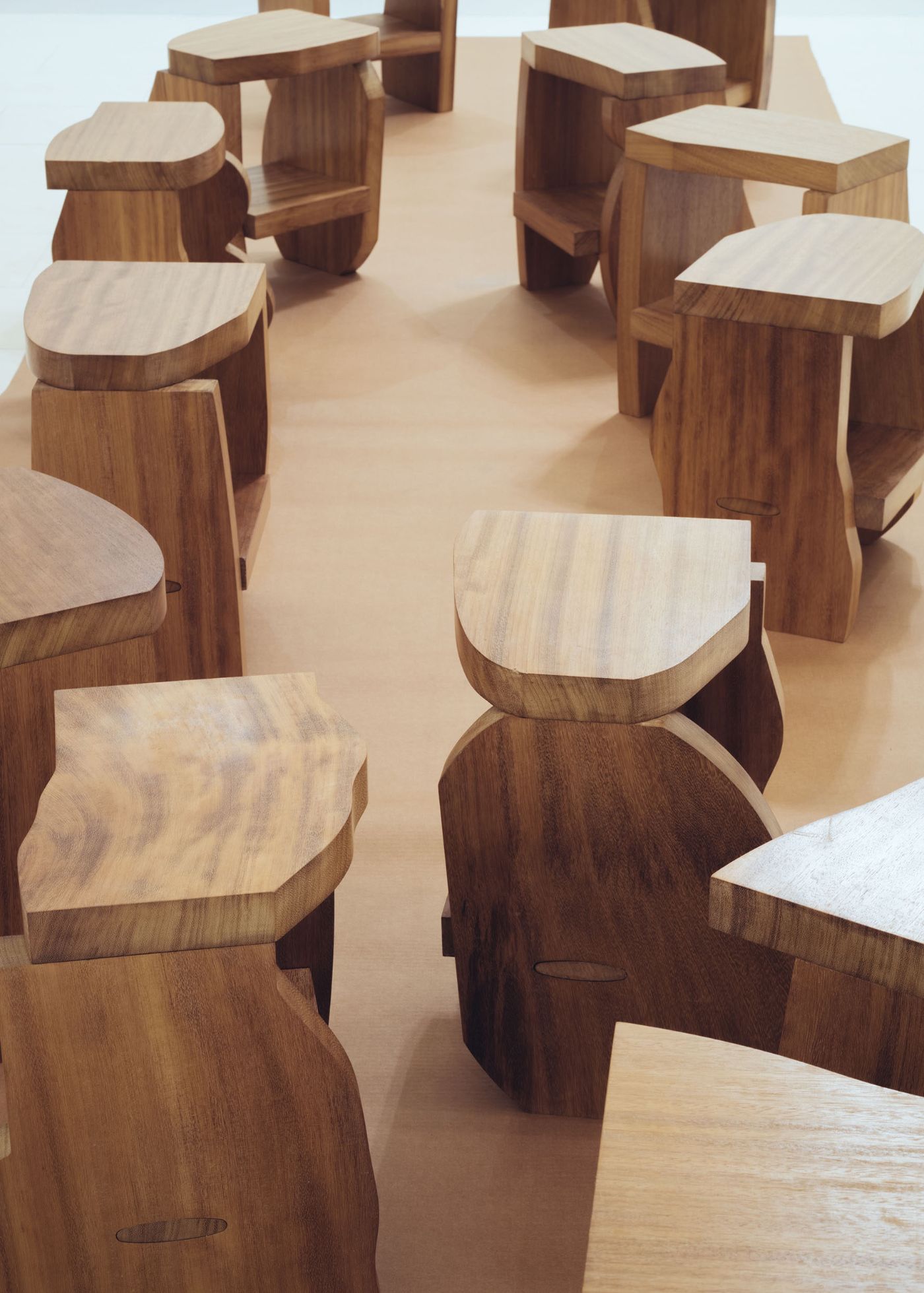 Selected Works by Bahraini-Danish — A collection of sculptural wooden stools arranged in a gallery setting, each featuring undulating tabletops with soft, rounded edges and organic cutout...