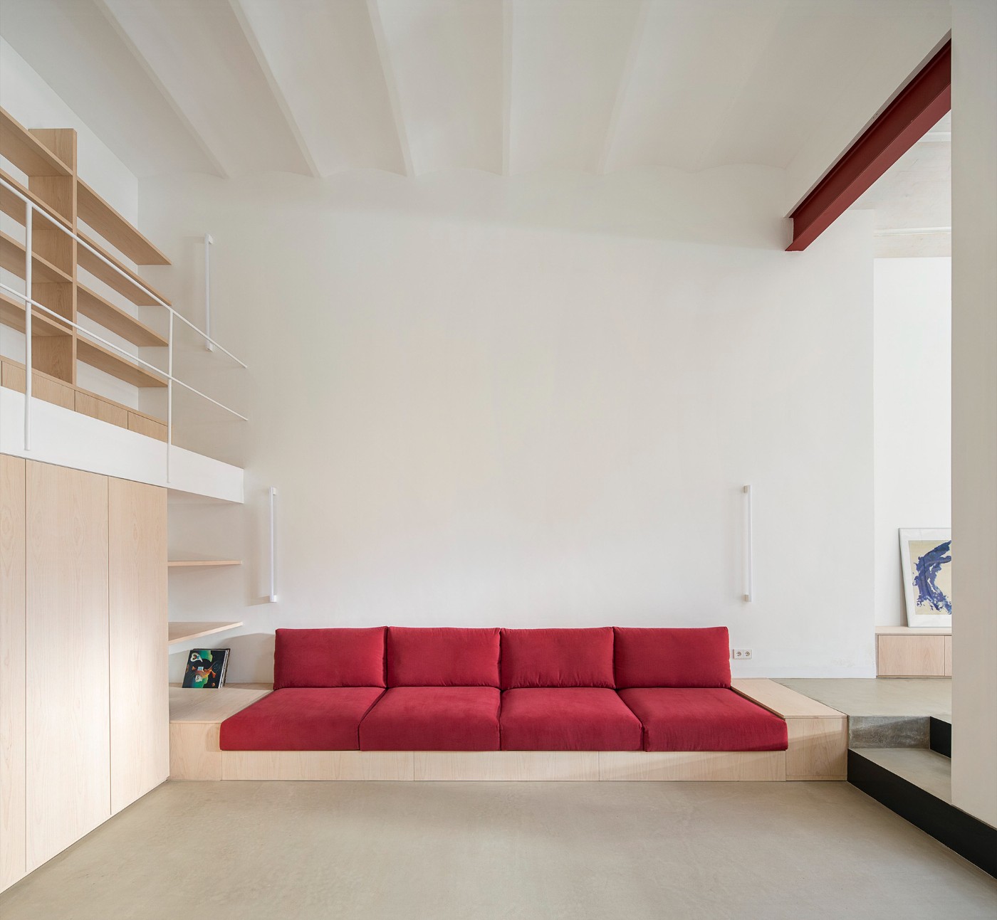 Red House by Aramé Studio — A single-story interior in Barcelona's converted Poblenou factory features a custom timber platform supporting a deep red upholstered four-part seating element.