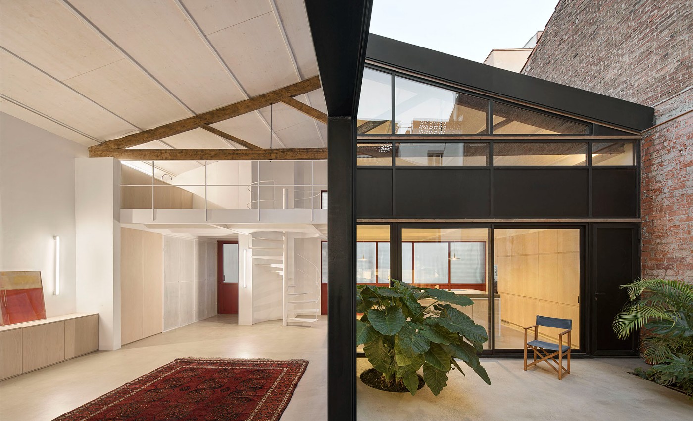 Red House by Aramé Studio — The courtyard view frames a double-height industrial space where exposed timber roof trusses with triangulated bracing span the full width, contrasting...