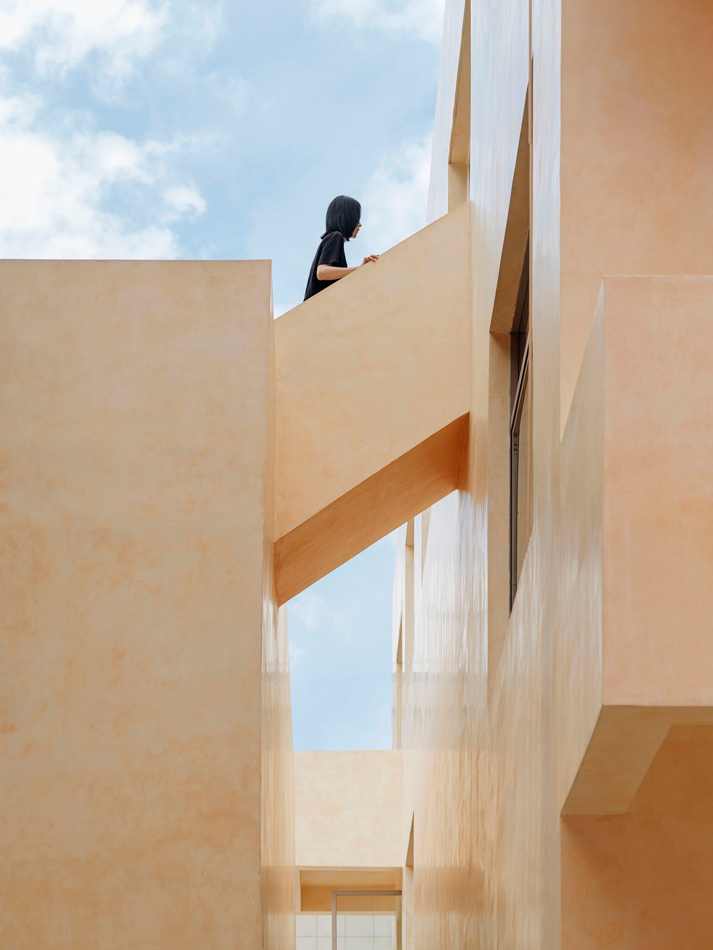 'The Field' by TEAM_BLDG by TEAM BLDG — A figure stands on a cantilevered concrete platform that extends horizontally across a multi-story void, framed by rendered walls in warm sandalwood-yellow...
