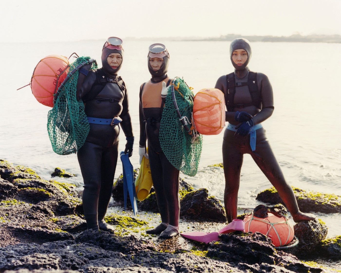 'The Last Mermaid' by Peter Ash Lee — Four women in black neoprene wetsuits stand on moss-covered volcanic rock at the water's edge, their diving equipment arranged around them.