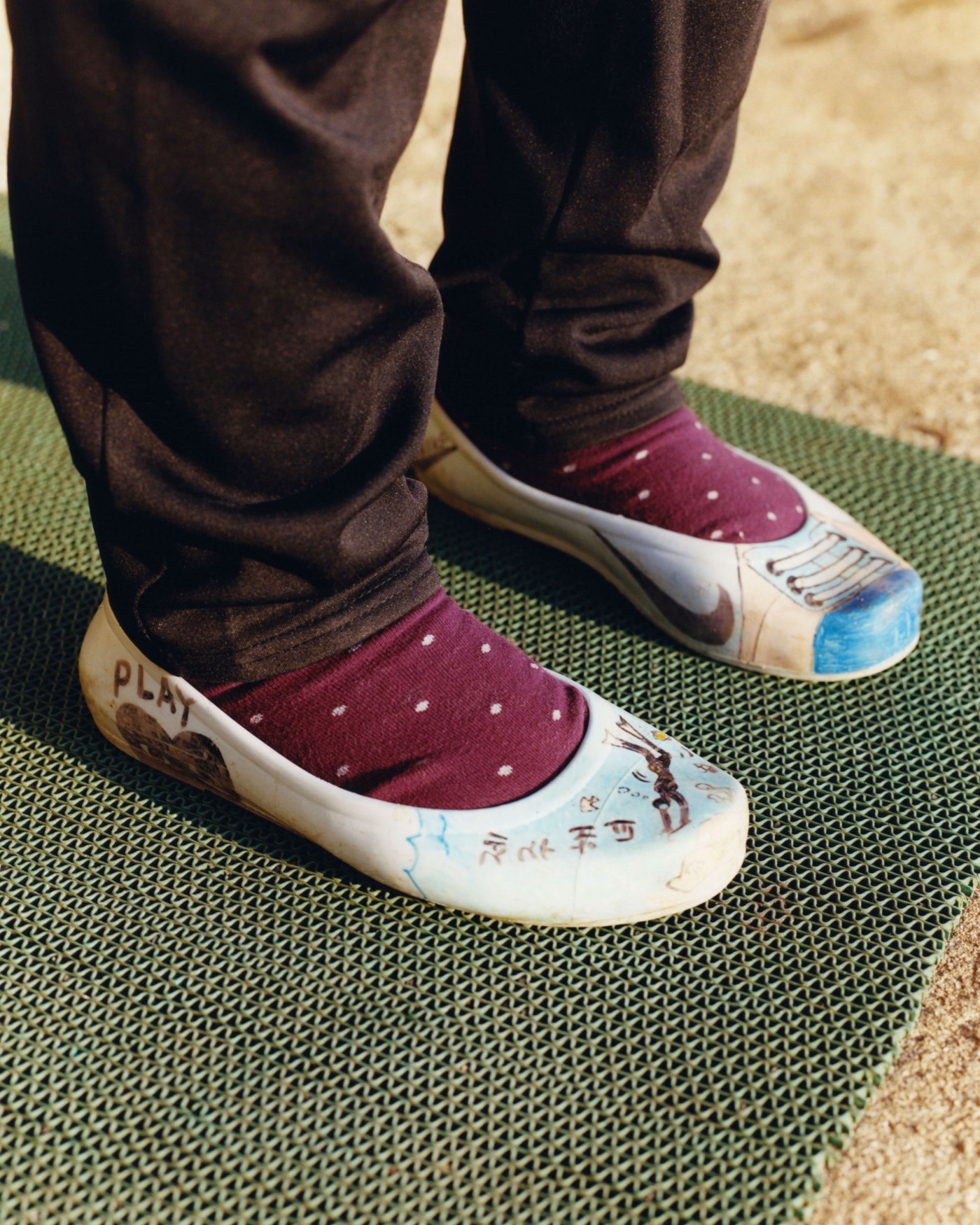 'The Last Mermaid' by Peter Ash Lee — A close-up detail shot capturing the lower legs and feet of a diver wearing cream canvas slip-on shoes with hand-painted blue and rust iconography on the...