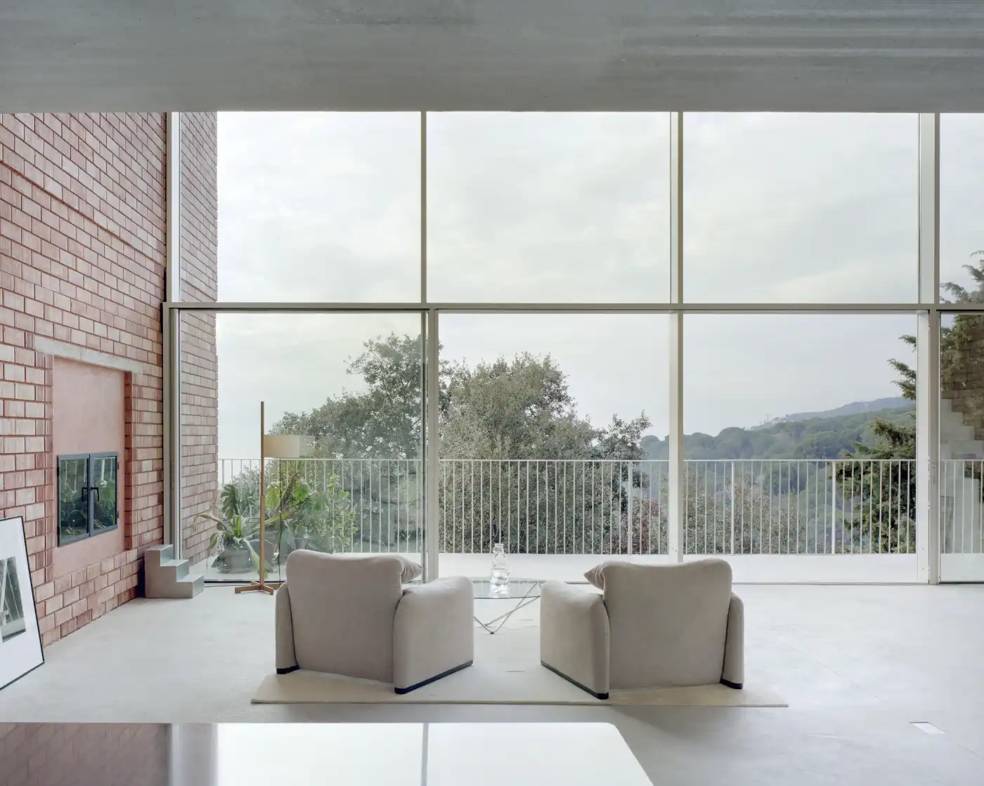 Casa Dosmurs by MESURA — Upper-floor seating area framed by extensive horizontal glazing with bronze-toned mullions, opening onto a metal-railed terrace overlooking densely...