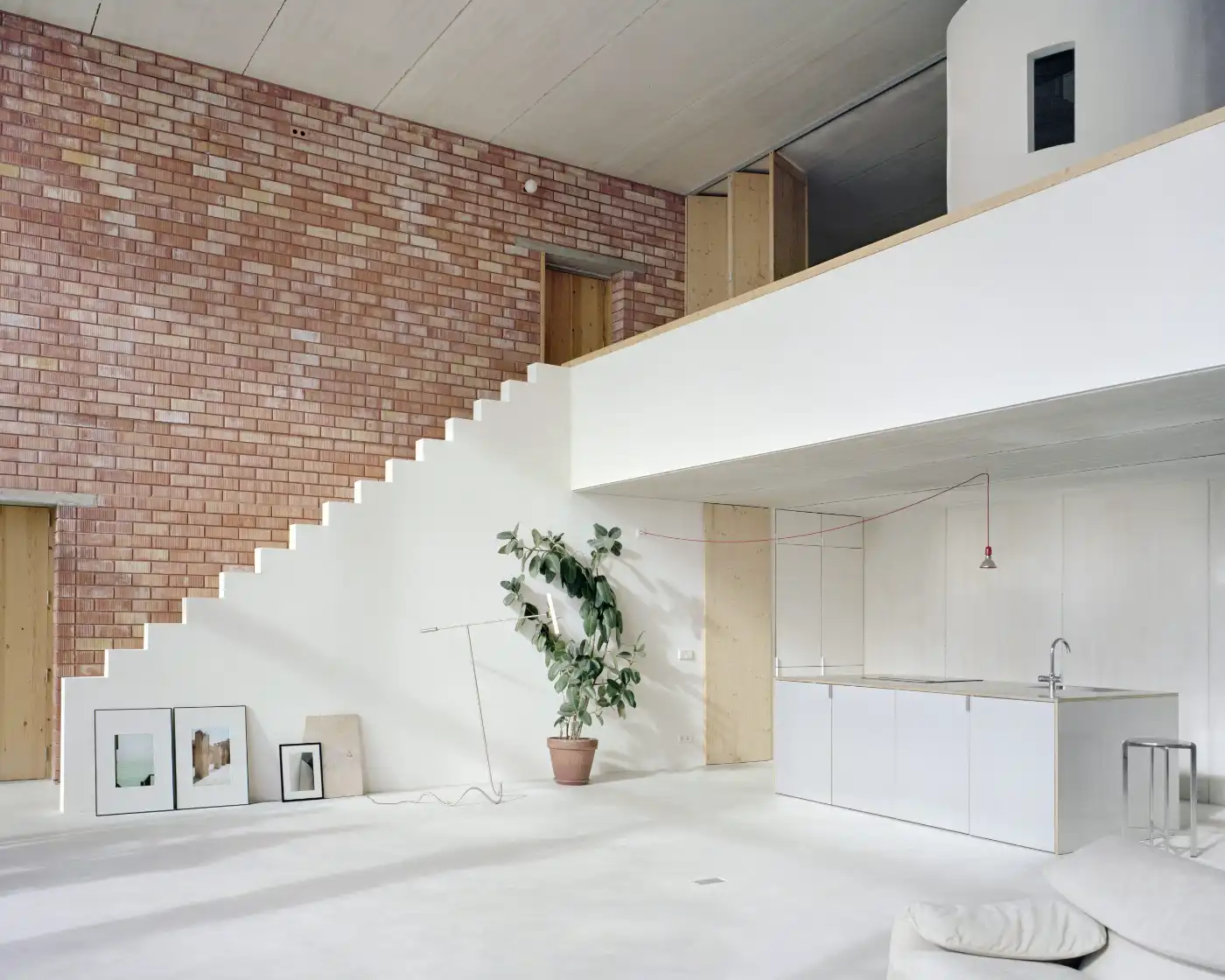 Casa Dosmurs by MESURA — Interior view of Casa Dosmurs showcasing the double-height living space with exposed red-brick upper wall contrasting against white render below.