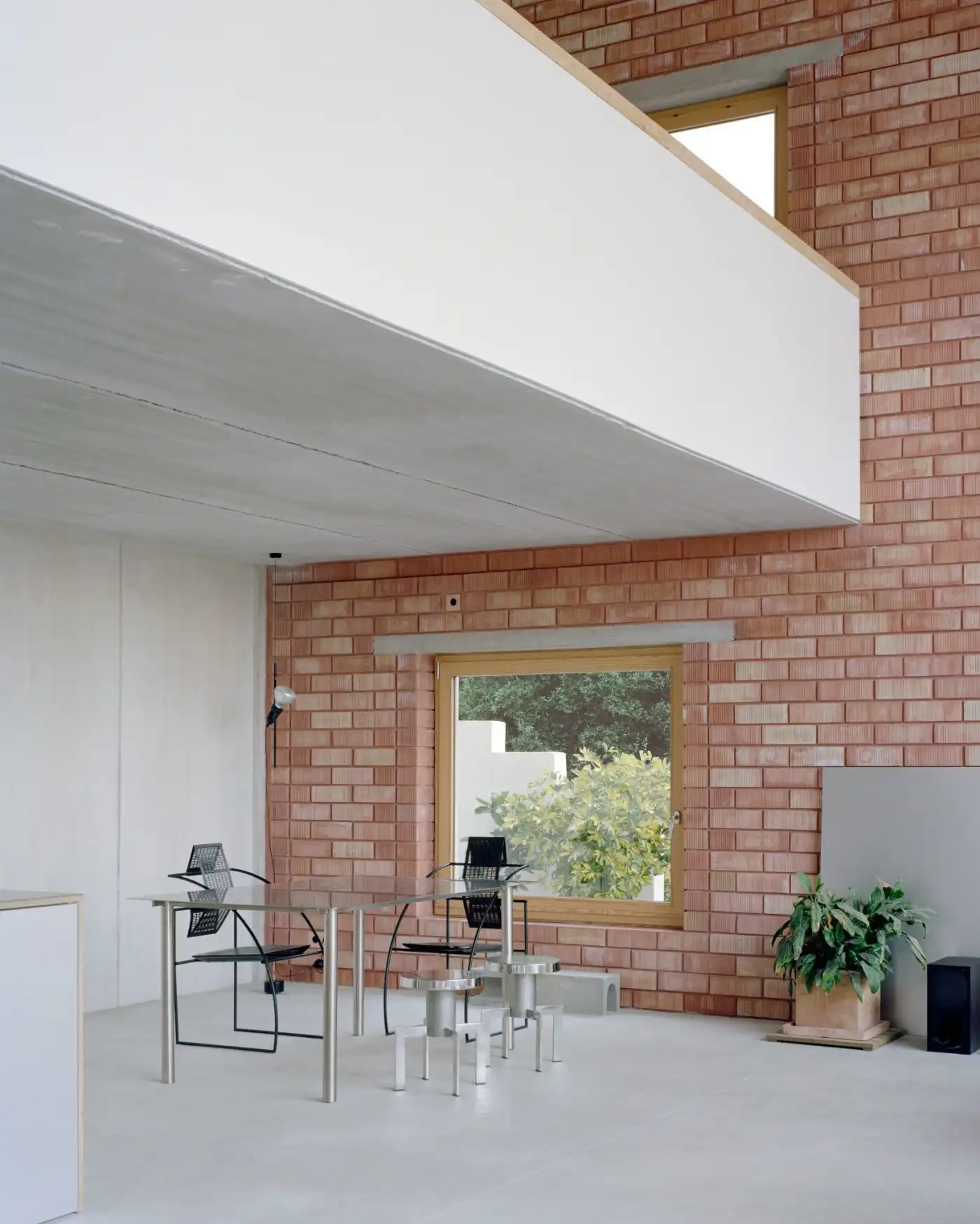 Casa Dosmurs by MESURA — A minimalist interior workspace carved into brick masonry, anchored by a large golden-bronze framed window opening onto vegetation.