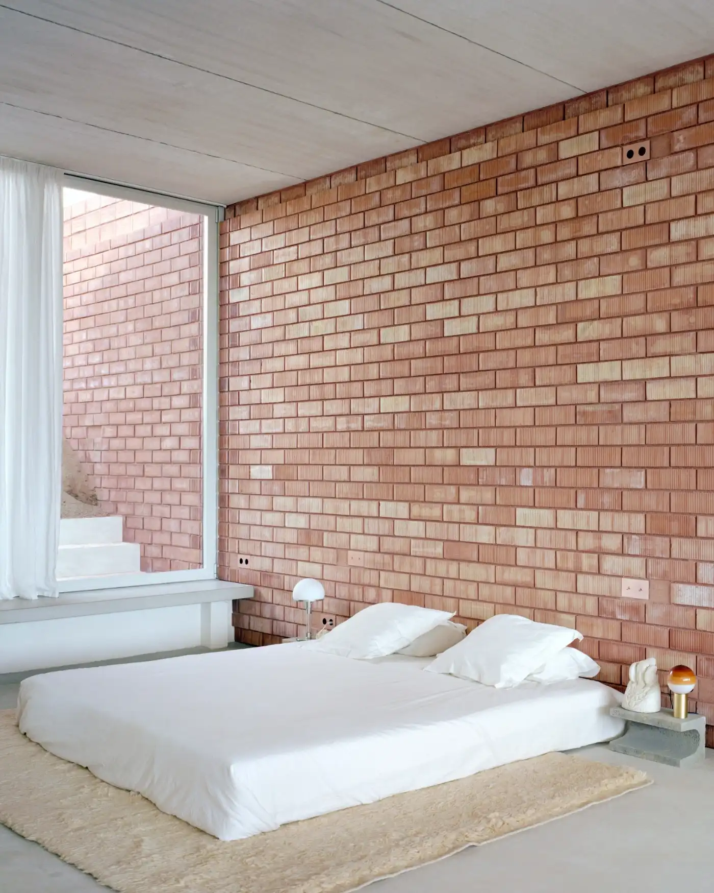 Casa Dosmurs by MESURA — Interior bedroom at Casa Dosmurs featuring a minimalist platform bed with white linens positioned against a full-height brick wall.