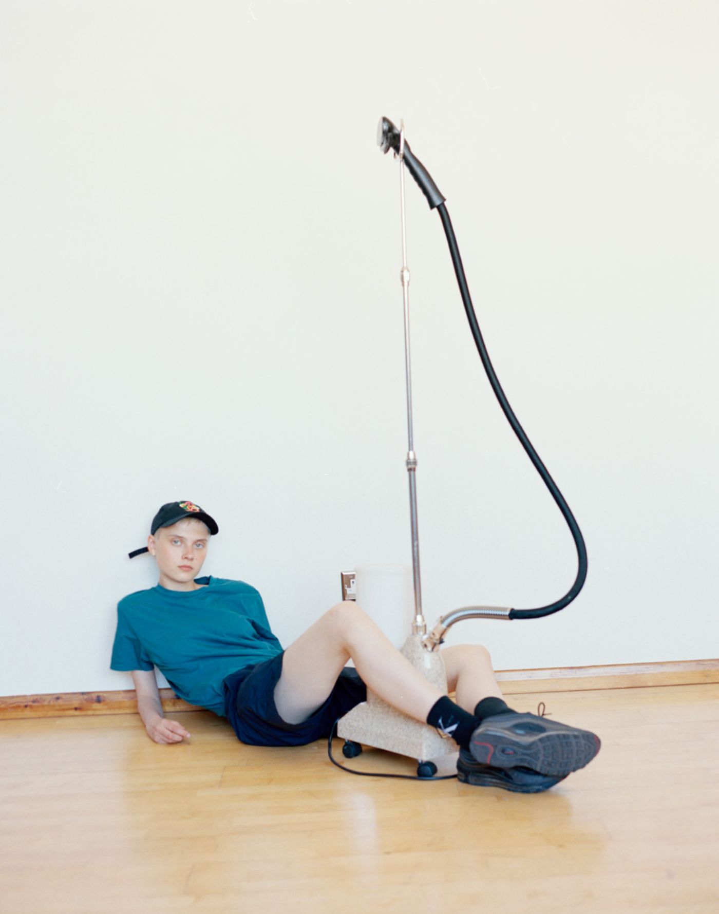 Visitors+ by Piczo — A young subject reclines on a wooden floor wearing a teal t-shirt and navy shorts, positioned beneath a pulley system mounted to the wall.