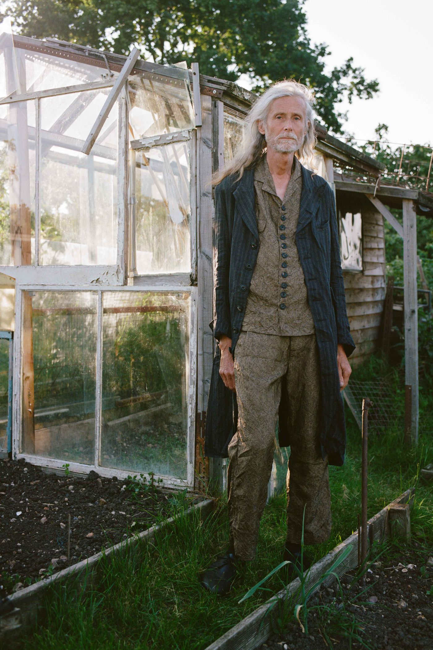 Spring Collection 2024 by John Skelton — A figure in layered wool and linen garments stands before a weathered timber-frame greenhouse structure in an active garden setting.