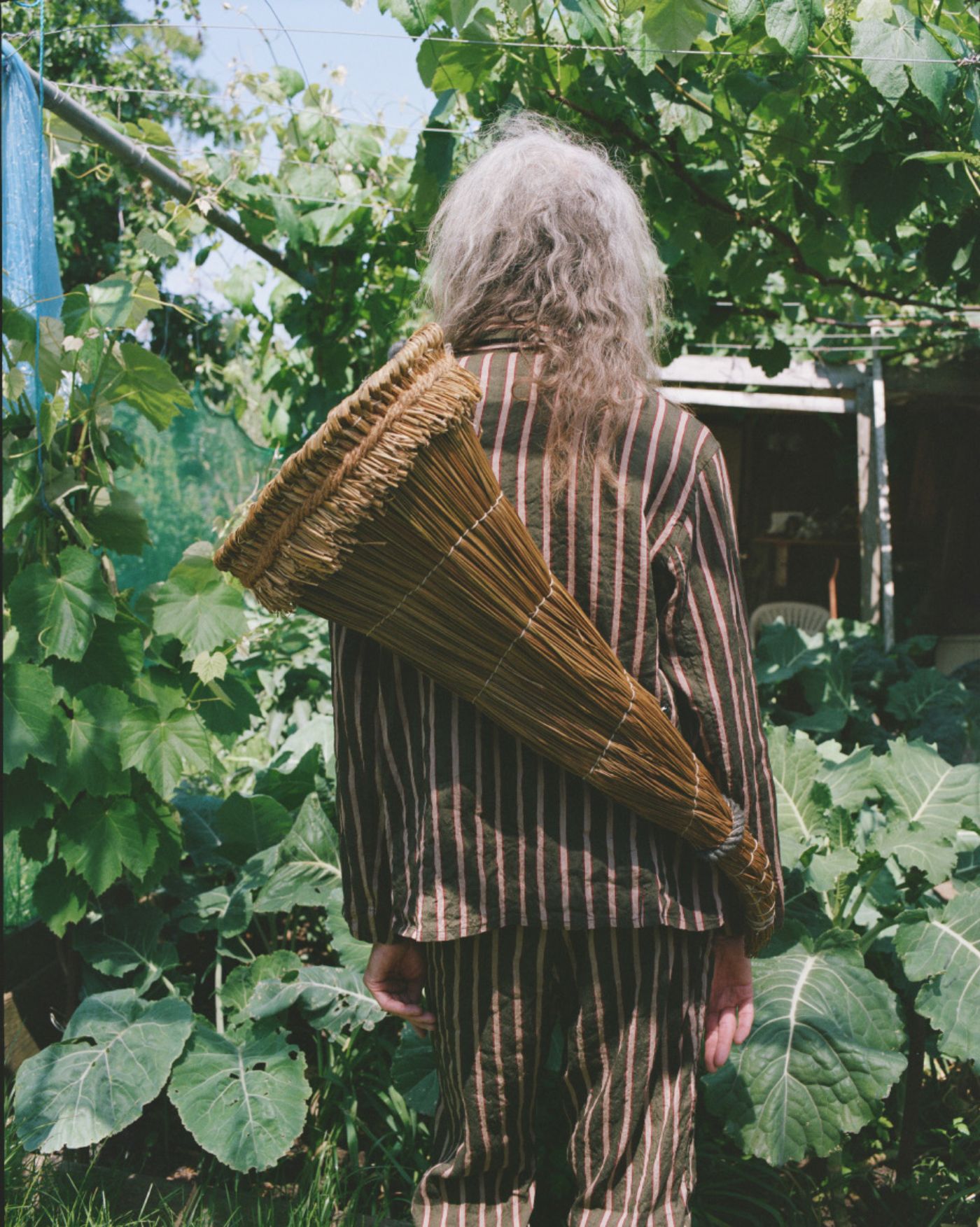 Spring Collection 2024 by John Skelton — A figure wearing a pinstriped wool jacket layered over an embroidered linen vest stands amid cultivated garden beds, carrying a woven wicker basket across...