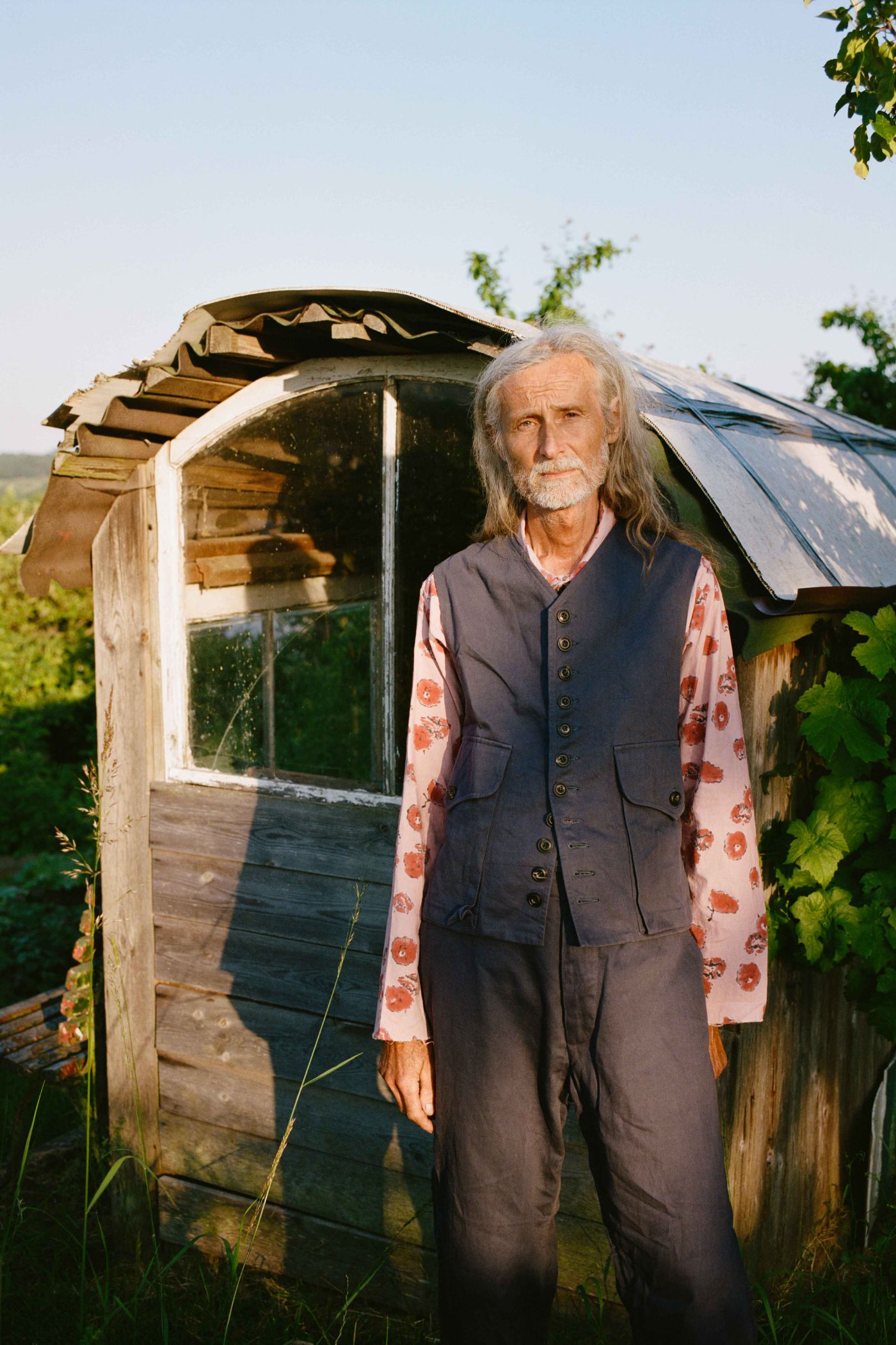Spring Collection 2024 by John Skelton — A figure stands before a weathered wooden garden shed with pitched roof and glazed door, positioned within cultivated allotment beds.