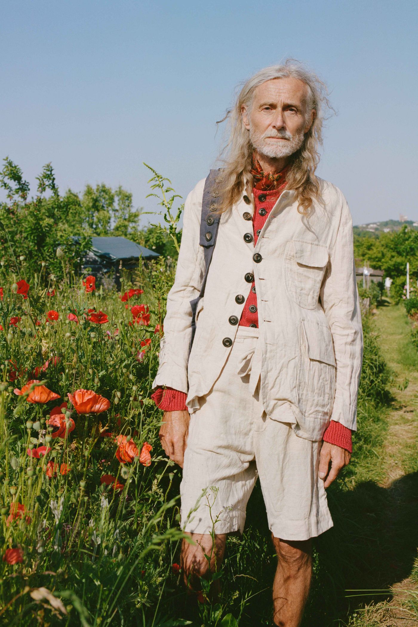 Spring Collection 2024 by John Skelton — A figure stands within a flourishing allotment garden, wearing a cream linen coat layered over a hand block-printed red waistcoat with embroidered detailing.