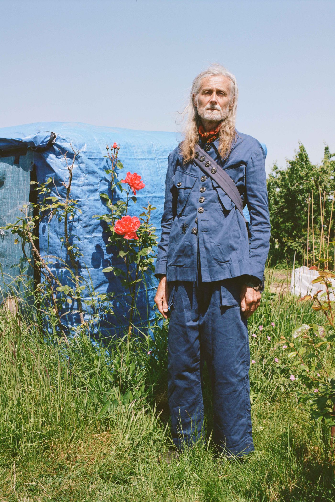 Spring Collection 2024 by John Skelton — A figure stands centered in an overgrown garden plot wearing a double-breasted navy linen jacket layered over an embroidered linen vest with hand...