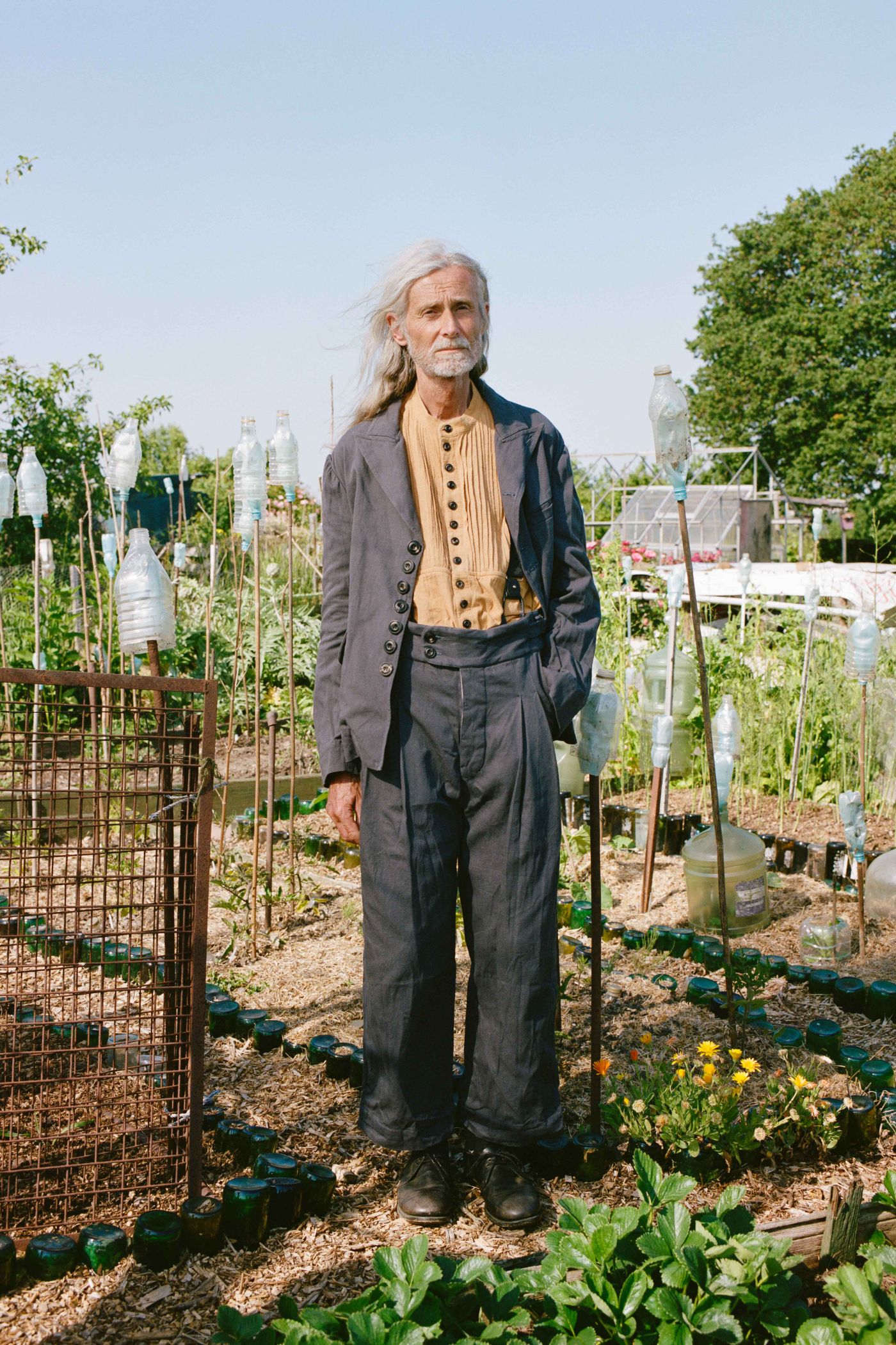 Spring Collection 2024 by John Skelton — A figure stands centered in a productive garden, wearing a layered ensemble of charcoal wool jacket and hand-embroidered cream linen vest with button detailing.