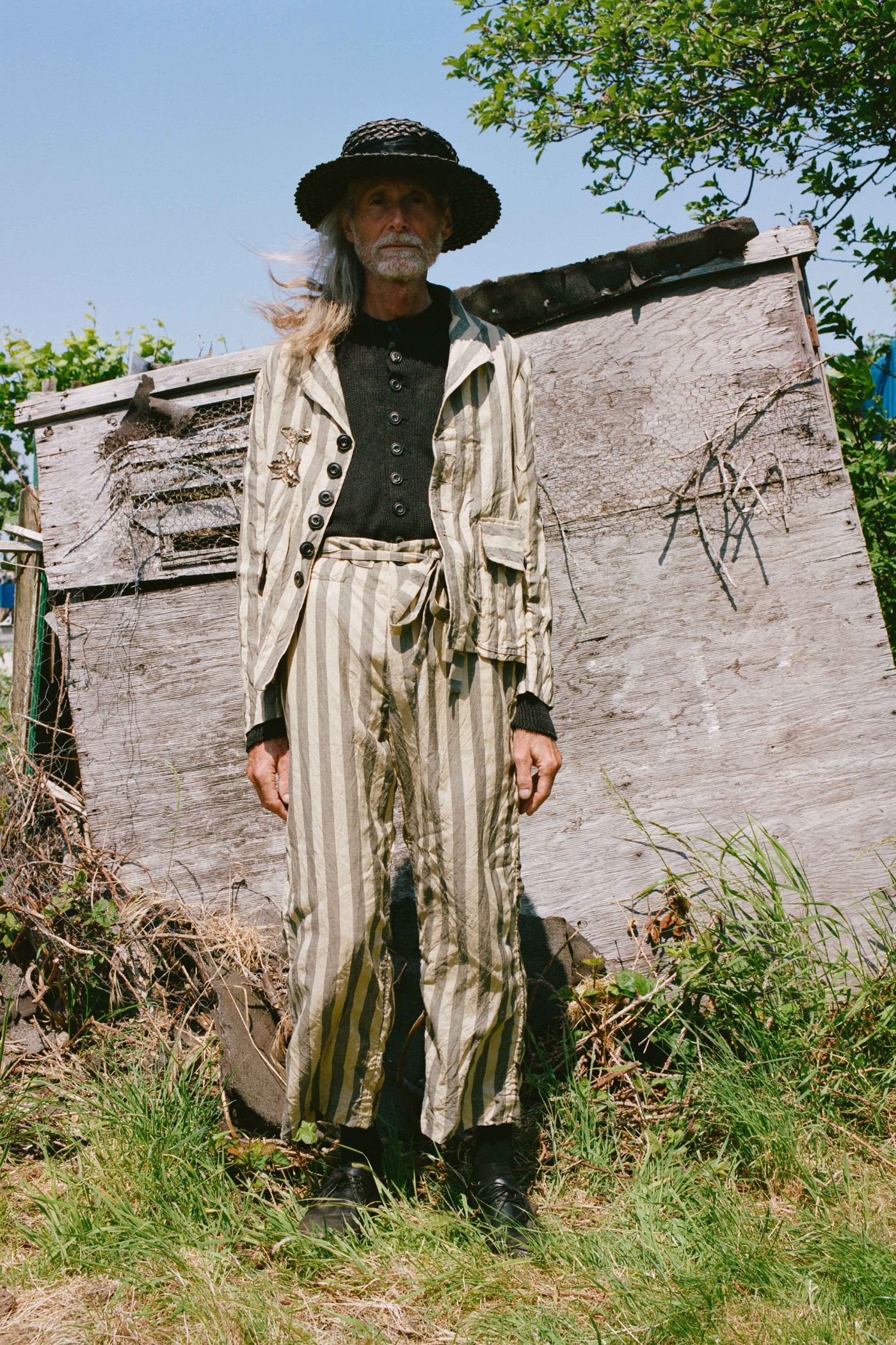 Spring Collection 2024 by John Skelton — A figure stands before weathered wooden fencing in garden grounds, wearing a layered ensemble from the Spring Collection 2024.