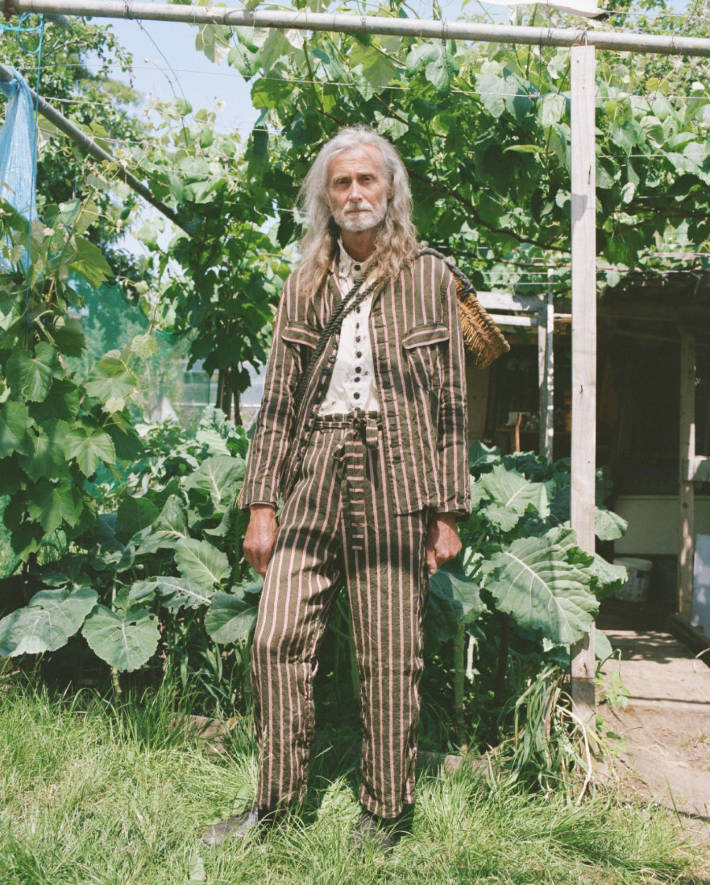 Spring Collection 2024 by John Skelton — A figure in a pinstriped wool jacket layered over a hand-embroidered linen vest stands within a productive garden enclosure.
