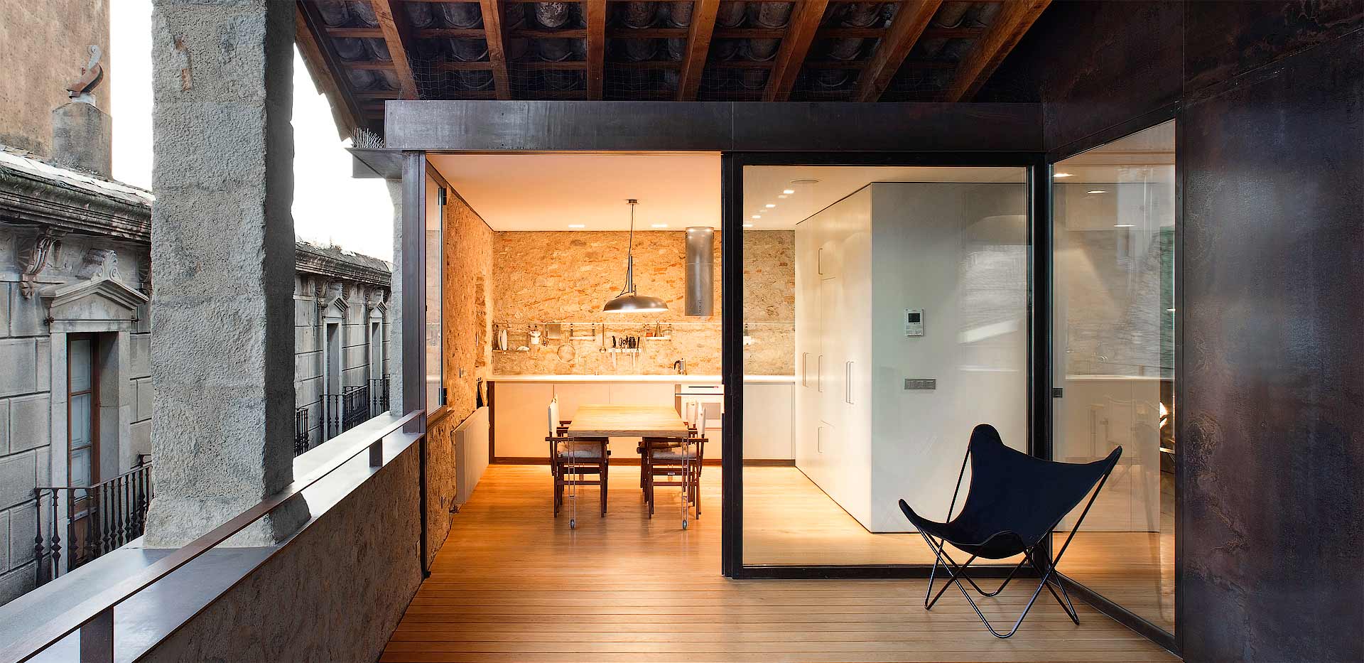 Alemenys 5 by Anna Noguera — A timber-decked terrace overlooks Girona's historic stone facades through frameless glass thresholds.