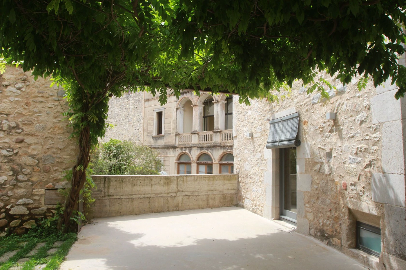 Alemenys 5 by Anna Noguera — A sunlit courtyard framed by ivy-draped stone walls reveals the restored Alemanys 5 façade beyond.