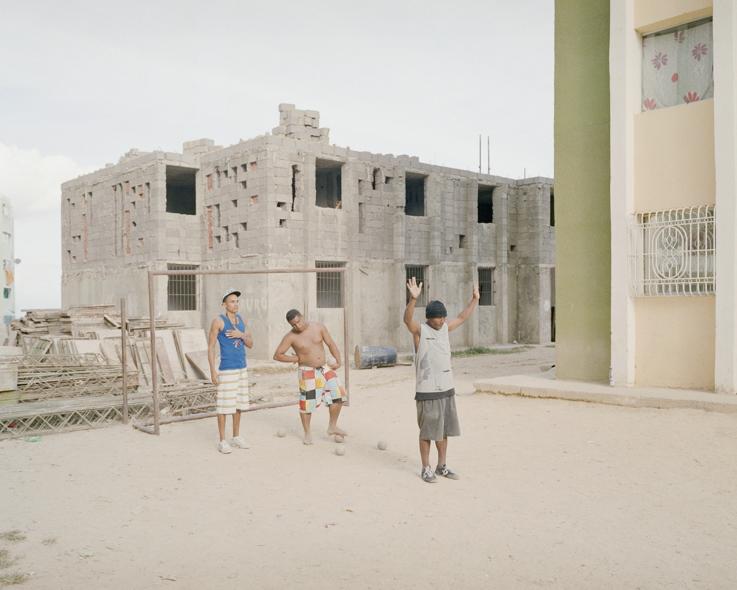 Buscando a Bolívar by Pietro Paolini — Three young boys play volleyball in a sandy courtyard bounded by an unfinished concrete-block structure with large window openings and visible rebar.