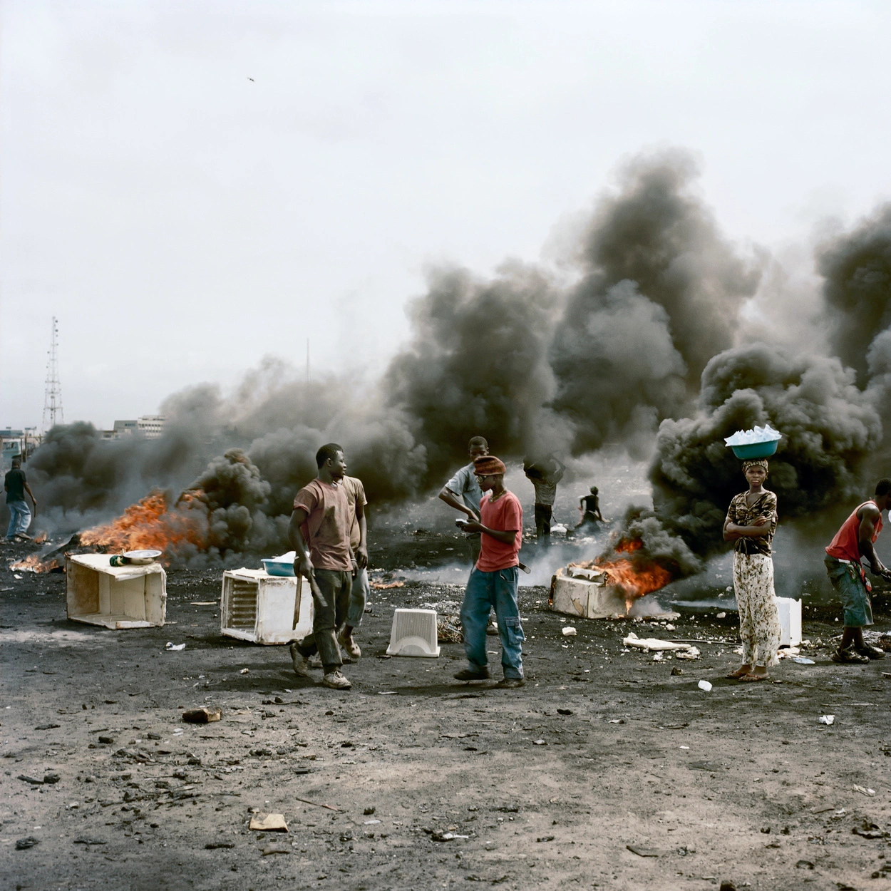Permanent Error by Pieter Hugo — Workers stand amid active burning waste at Agbogbloshie landfill, their bodies and clothing coated in grey ash and fine particulate matter.