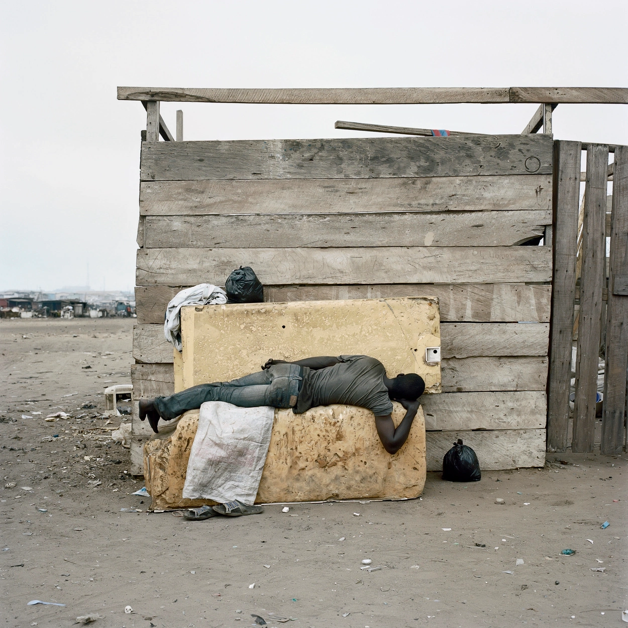 Permanent Error by Pieter Hugo — A figure lies prone across a rammed-earth block positioned against weathered timber structures in Accra's Agbogbloshie landfill.