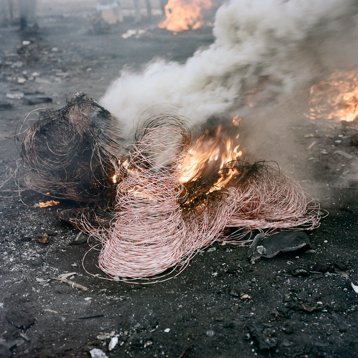 Permanent Error by Pieter Hugo — Coiled copper wire, actively burning with visible flame and white smoke, lies on dark asphalt ground scattered with debris.