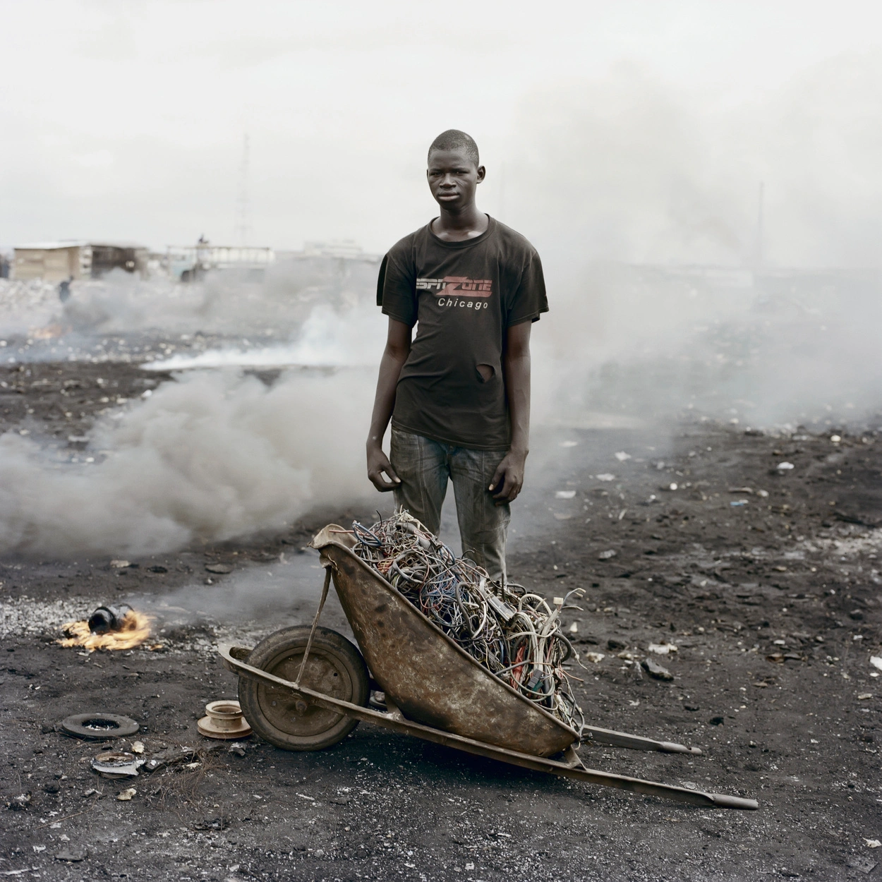 Permanent Error by Pieter Hugo — A young man stands centered in Agbogbloshie's landfill, wearing a black Chicago-branded t-shirt dusted with ash and fine particulate matter.