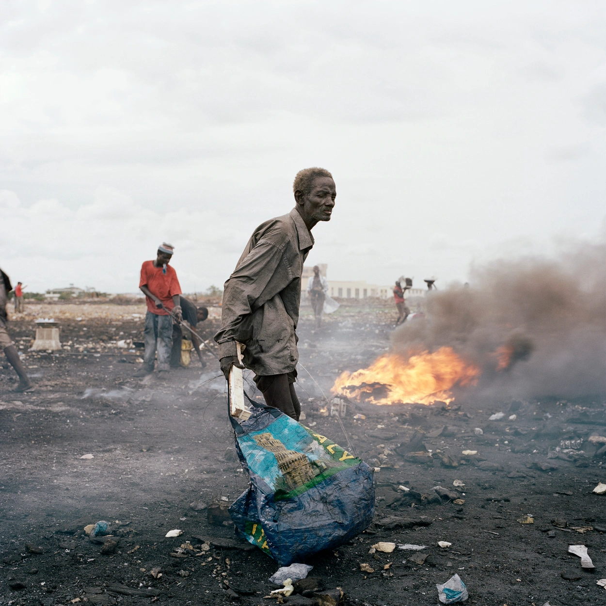 Permanent Error by Pieter Hugo — A worker stands in Agbogbloshie landfill holding a discarded plastic bag printed with architectural imagery, his dark skin and clothing visibly coated in...