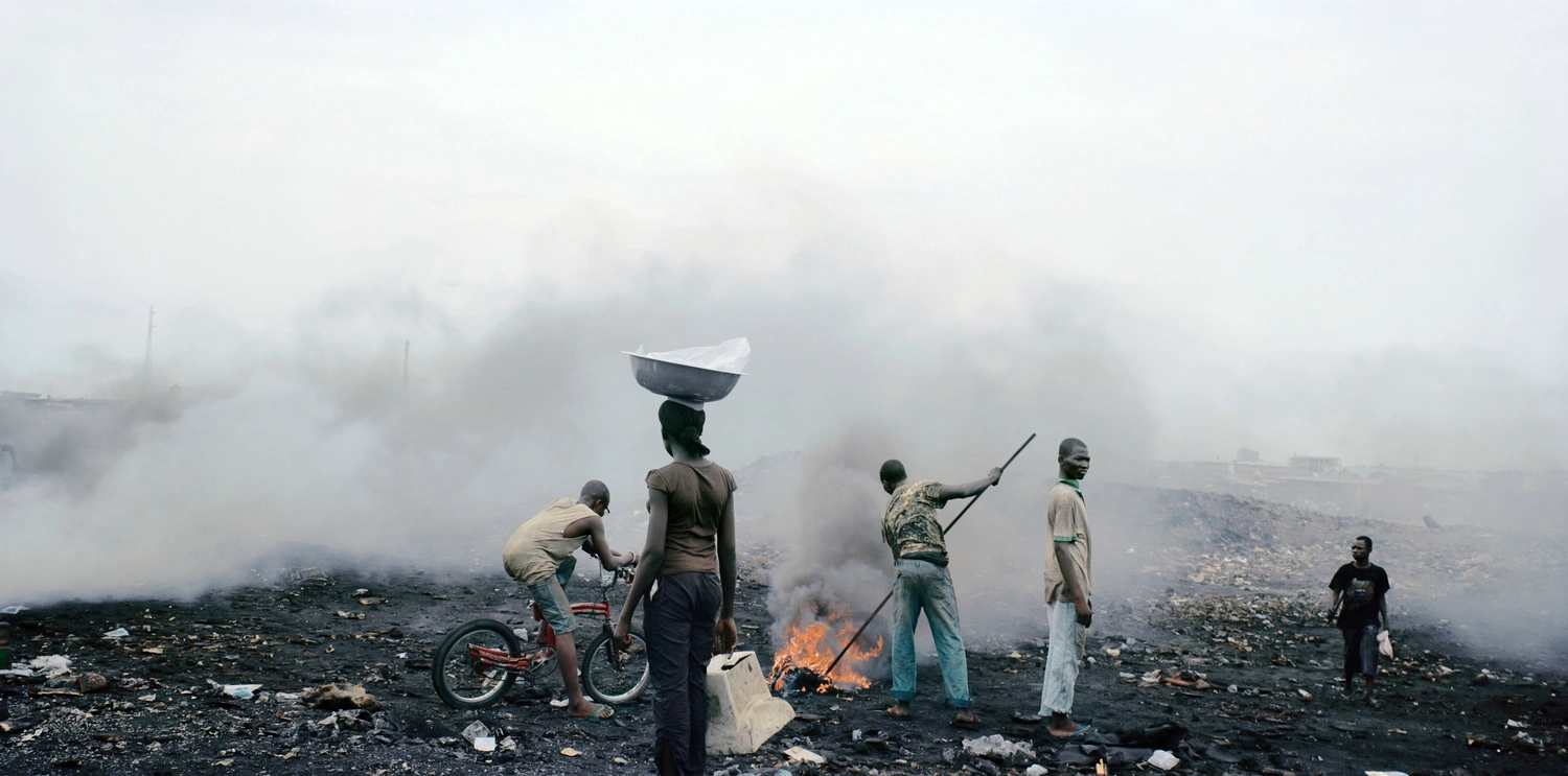 Permanent Error by Pieter Hugo — Five figures occupy a blackened landfill scattered with waste, dense smoke and ash obscuring the background.