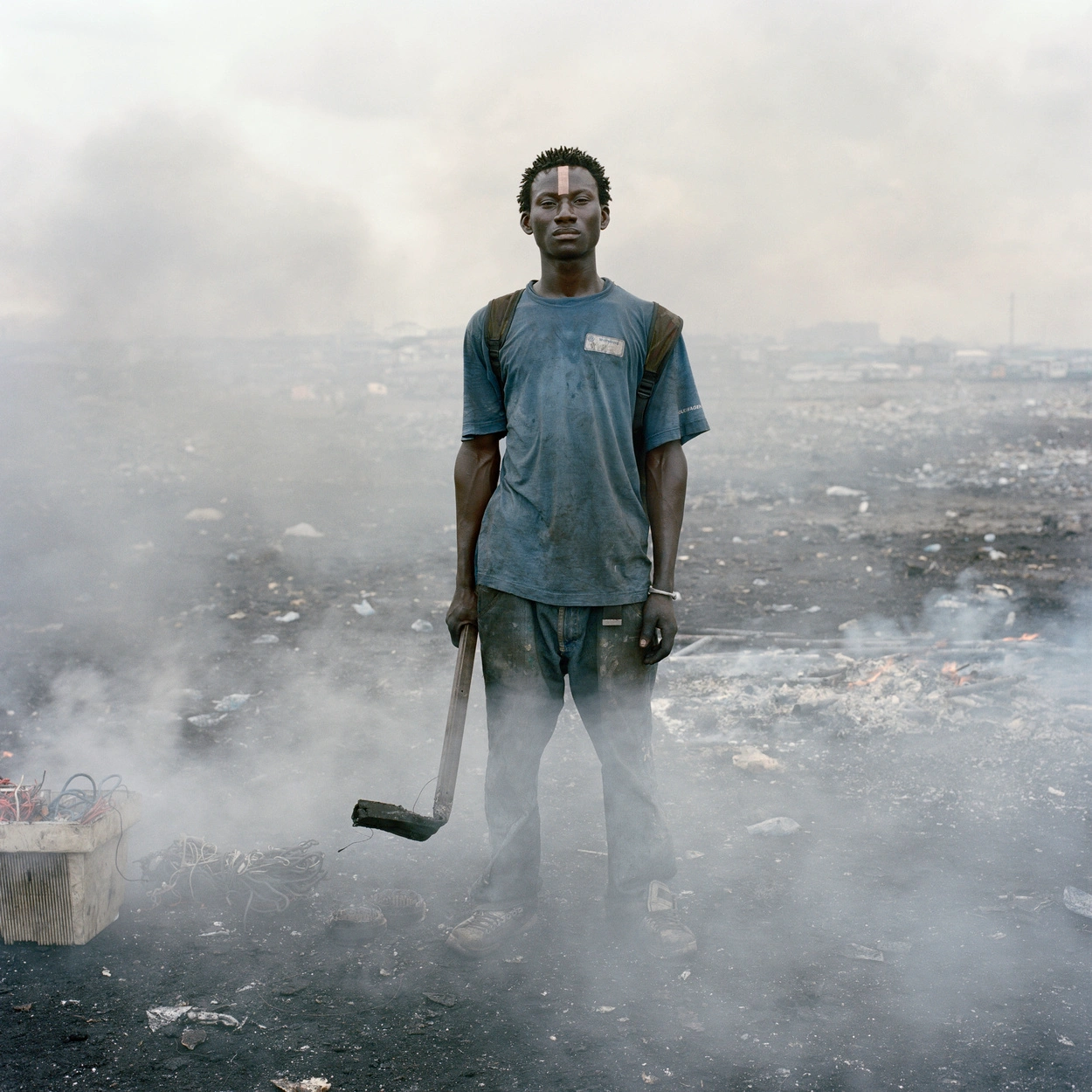 Permanent Error by Pieter Hugo — A young man stands centered in the frame against a smoke-obscured landscape, his dark skin coated in fine ash particulate matter.