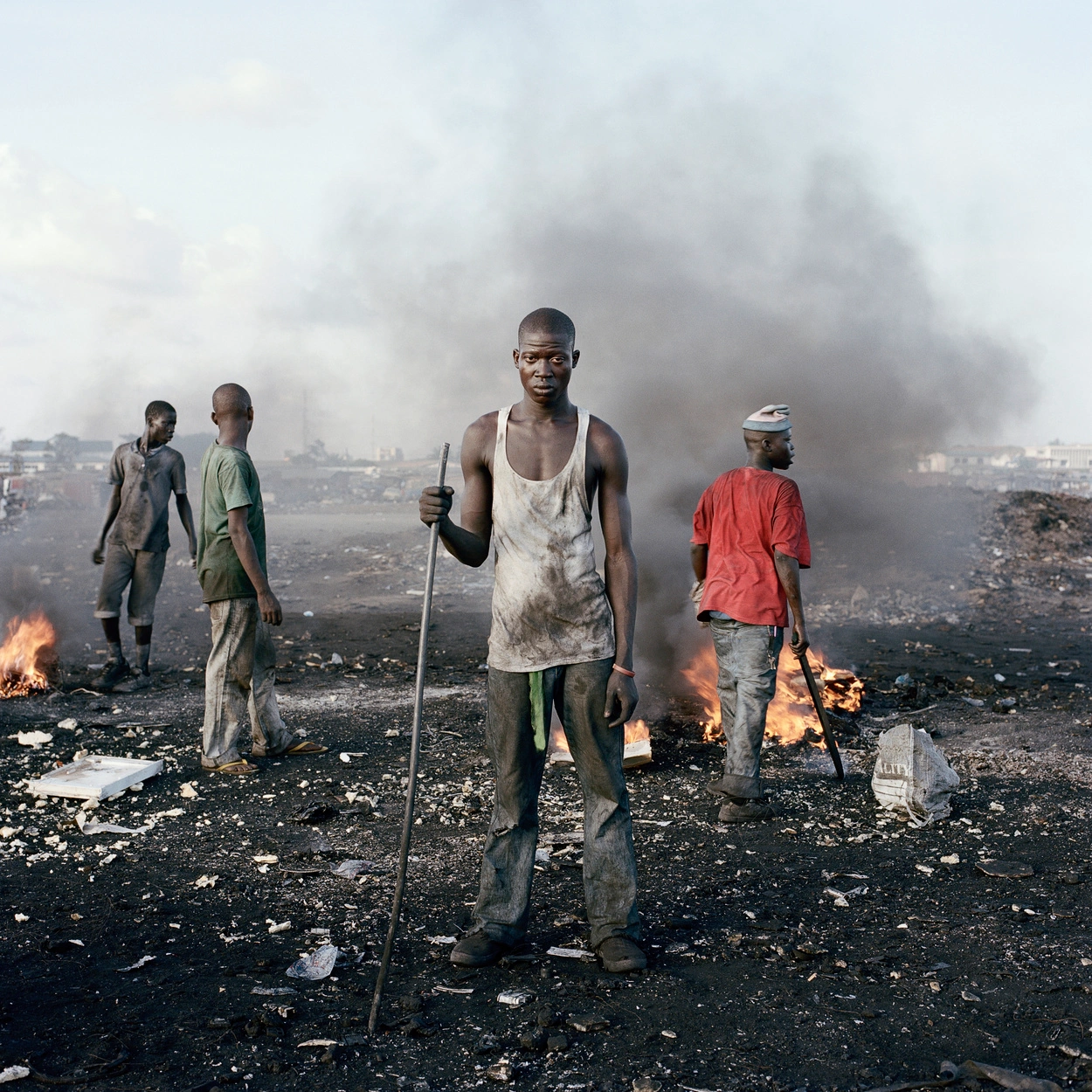 Permanent Error by Pieter Hugo — A young man stands centered in the frame at Agbogbloshie landfill, his white tank top and exposed skin heavily coated with grey ash and fine particulate...