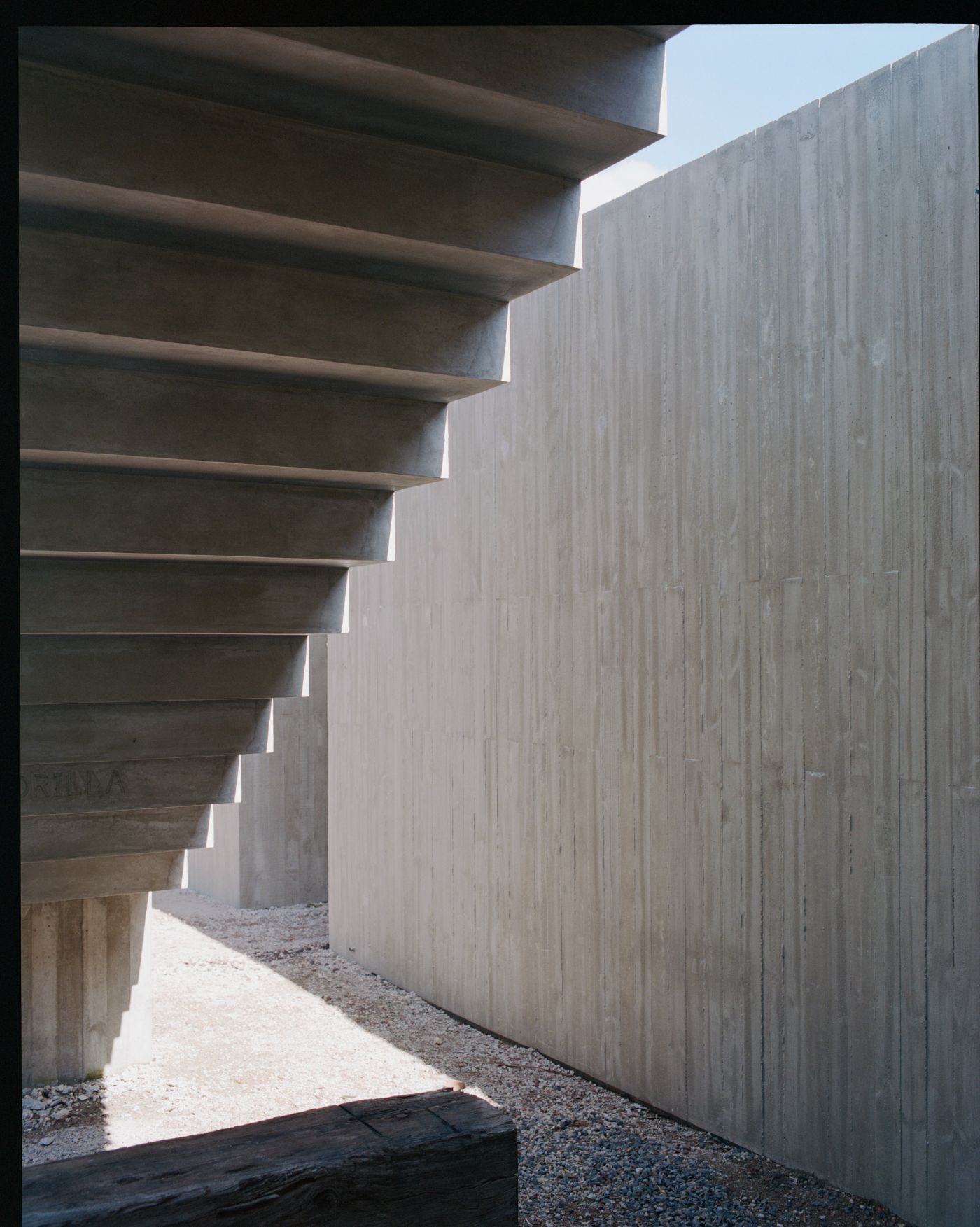 Mori House by Aires Mateus + MAArchitects — A narrow passage frames a cantilever stair with board-formed concrete treads projecting from the left wall, their undersides casting sharp shadows.