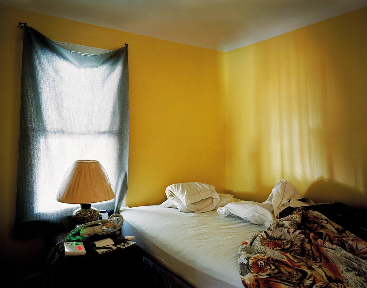 The Prospect of Immortality by Murray Ballard — A sparsely furnished bedroom in yellow ochre, shot in natural daylight from a window.