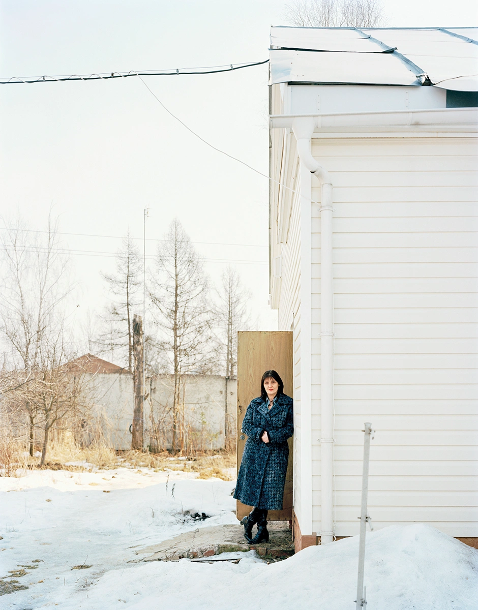 The Prospect of Immortality by Murray Ballard — A woman in a dark patterned coat stands at the threshold of a white corrugated metal structure in a winter landscape.