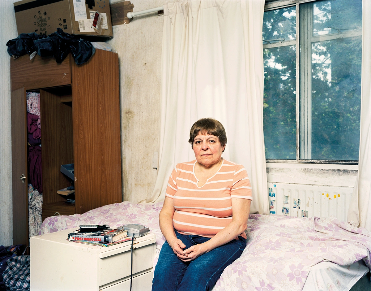 The Prospect of Immortality by Murray Ballard — A woman sits centered on a bed with lavender floral bedding in a modest bedroom, wearing an orange and white striped shirt and blue jeans.
