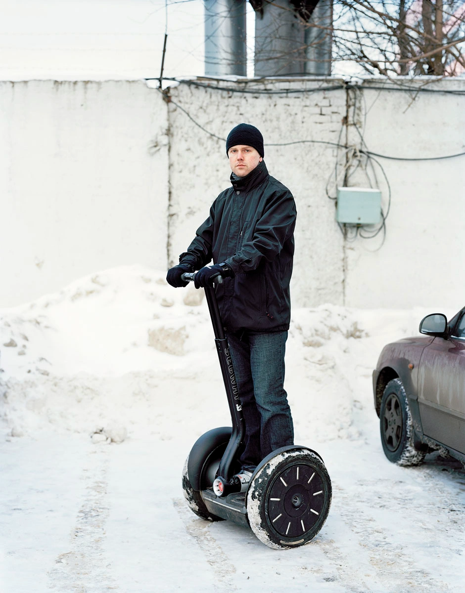 The Prospect of Immortality by Murray Ballard — A man stands centered on a Segway in a snow-covered industrial courtyard, wearing a black insulated jacket and knit beanie.