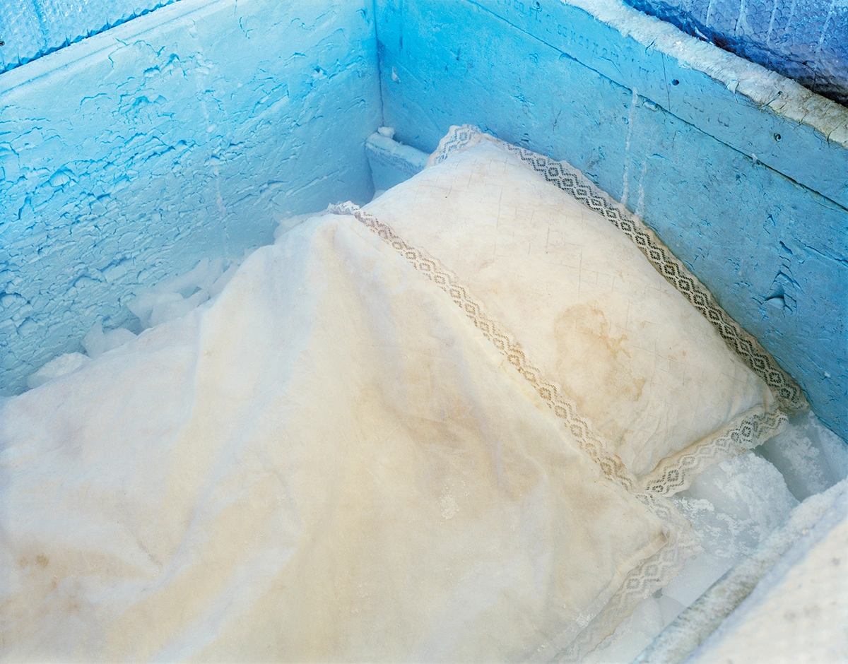 The Prospect of Immortality by Murray Ballard — A close-up view of textile bundles wrapped in cream linen with lace trim, arranged within a bright blue wooden crate.