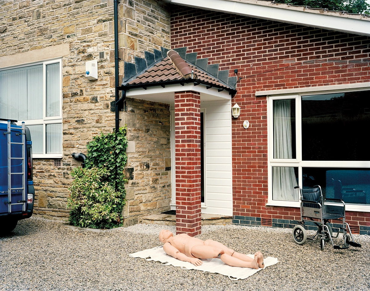 The Prospect of Immortality by Murray Ballard — A life-size anatomical mannequin lies supine on canvas in the gravel courtyard of a residential property, positioned centrally beneath a brick chimney stack.
