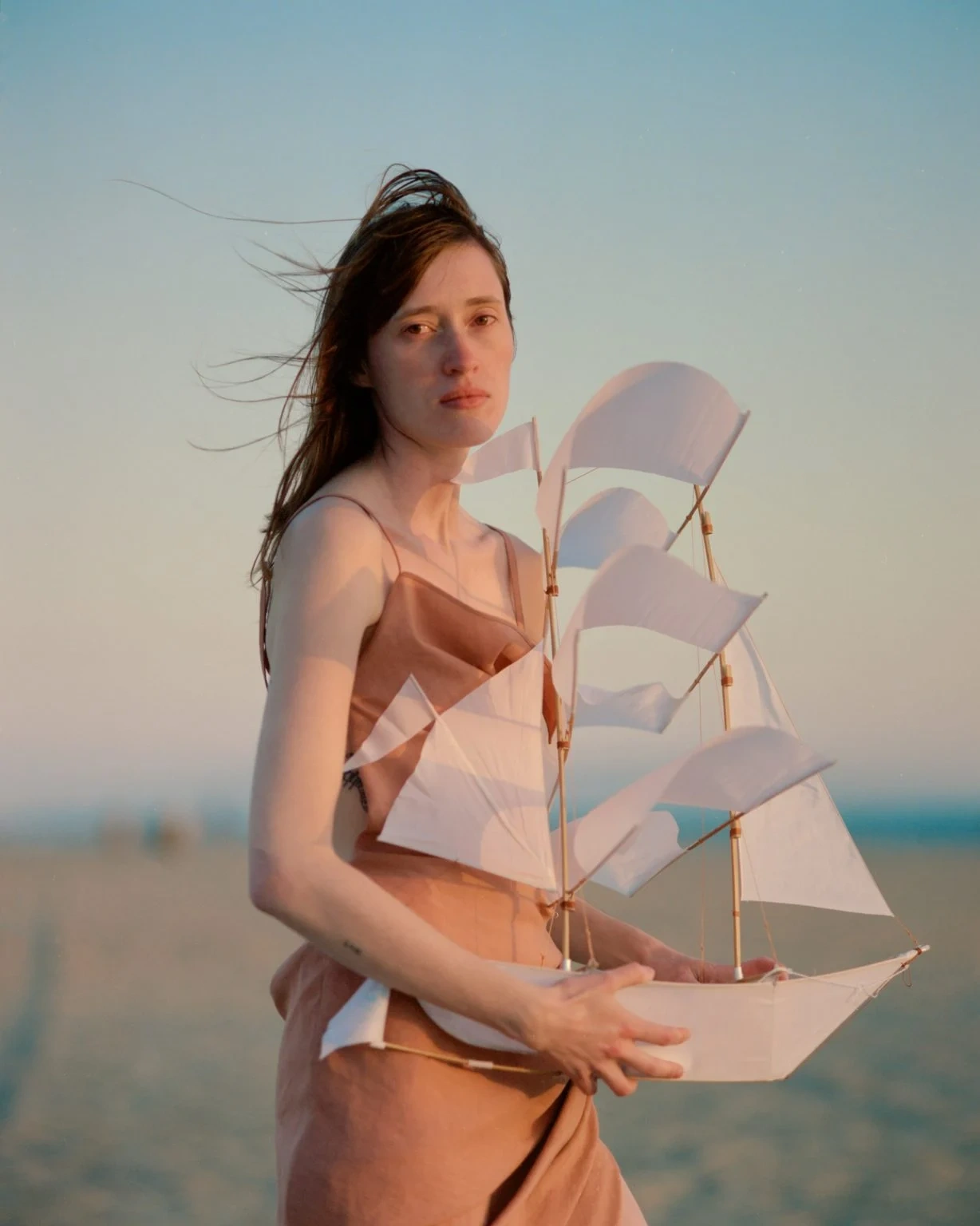 Selected works by Arianna Lago — A woman in a terracotta linen dress holds a delicate paper model sailing ship against a coastal backdrop.