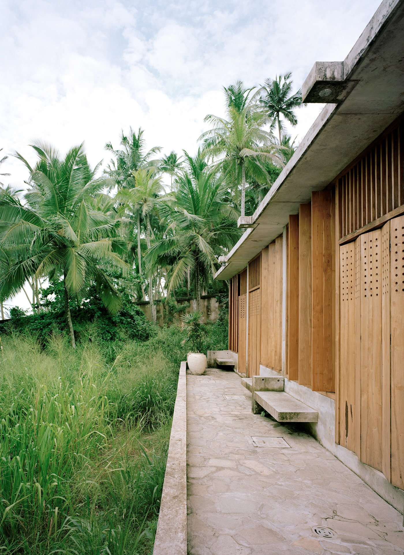 057 Guesthouse Hiriketiya by Atelier Abraha Achermann — A covered passage flanks the guesthouse's timber-shuttered facade, its weathered wood doors detailed with perforated geometric patterns.